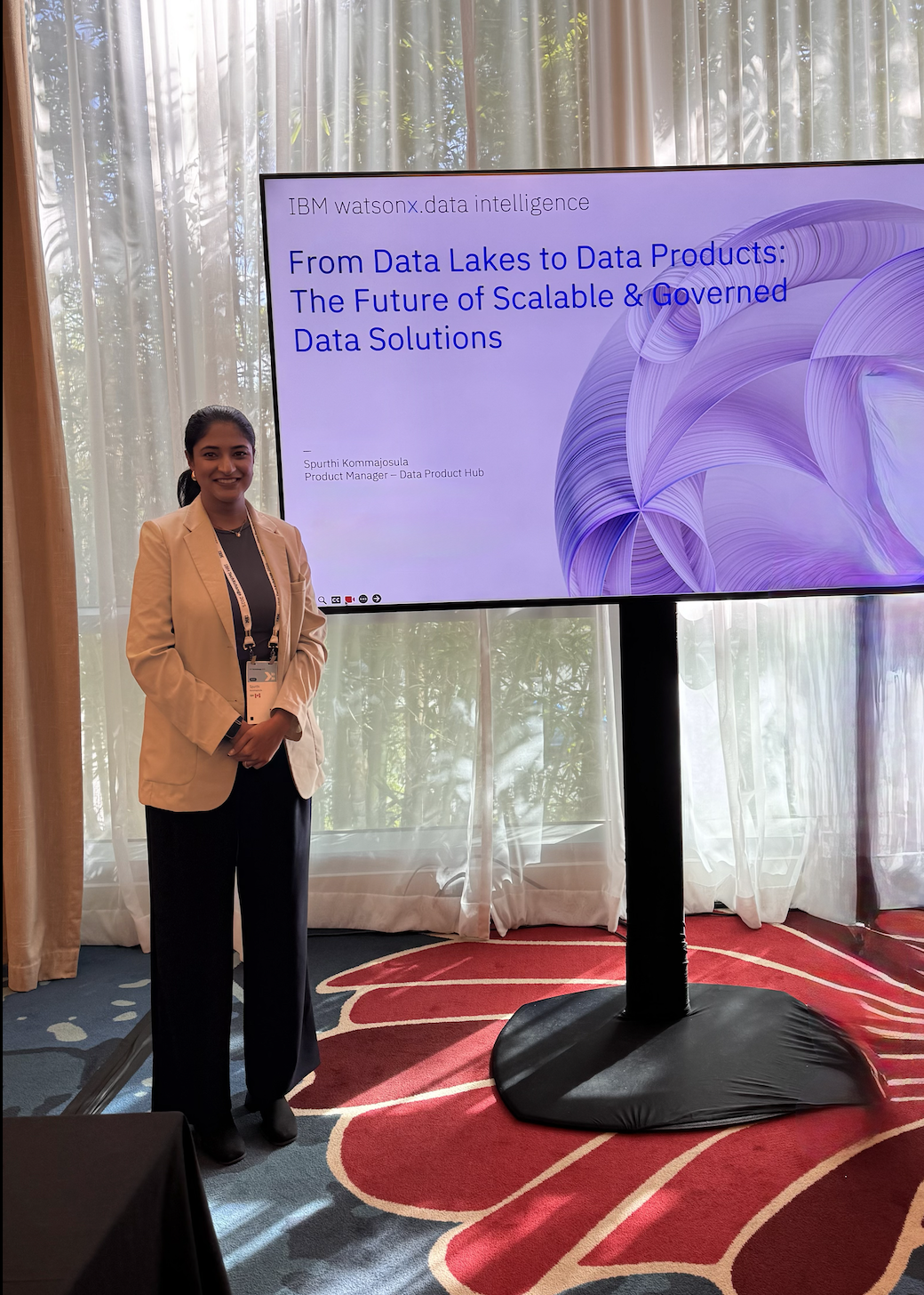 A woman in a beige blazer and dark pants standing next to a large screen with a presentation slide about data solutions, in a conference room with curtains and natural light.