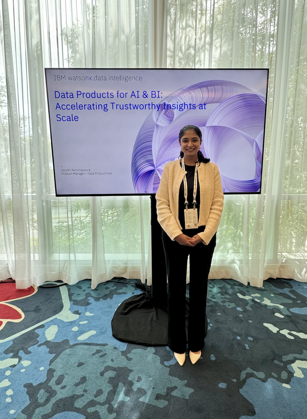A woman standing in front of a presentation screen at a conference, with a window with curtains behind her. The screen displays the IBM Watson logo and a presentation title about data products for AI and BI.