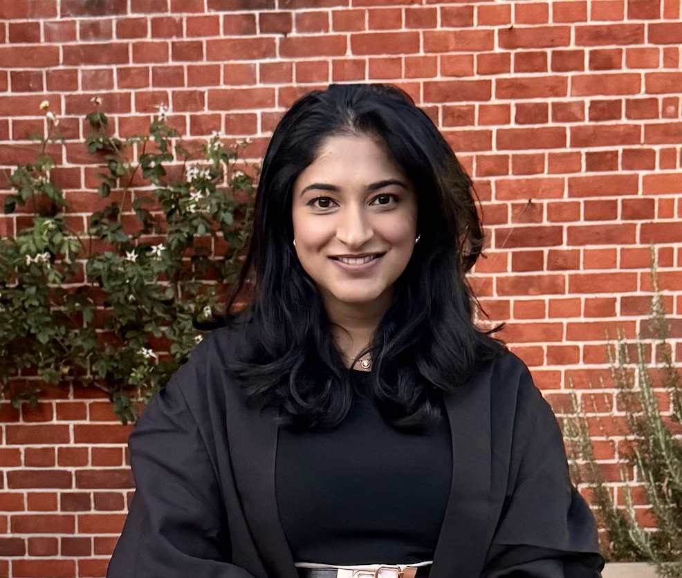 Young woman with black hair smiling, standing outdoors in front of a red brick wall with greenery.