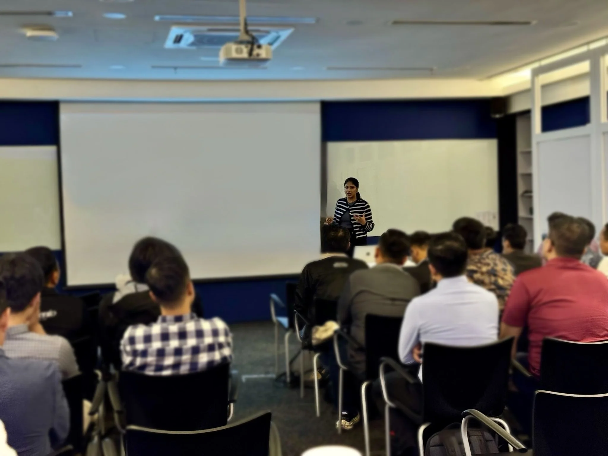 A woman giving a presentation to a group of people in a classroom or conference room. The room has a large whiteboard and a projector mounted on the ceiling, with attendees seated and listening.