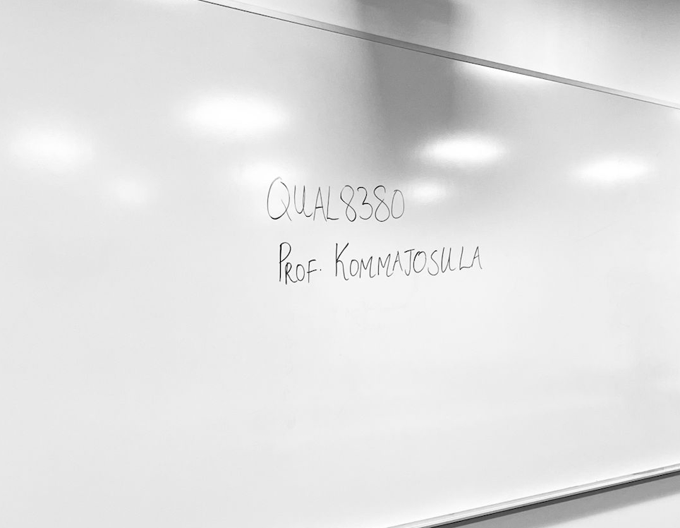 Whiteboard with handwritten text: 'QUAL 8380' and 'Prof. Kommatosulla' in a classroom or lecture hall setting.