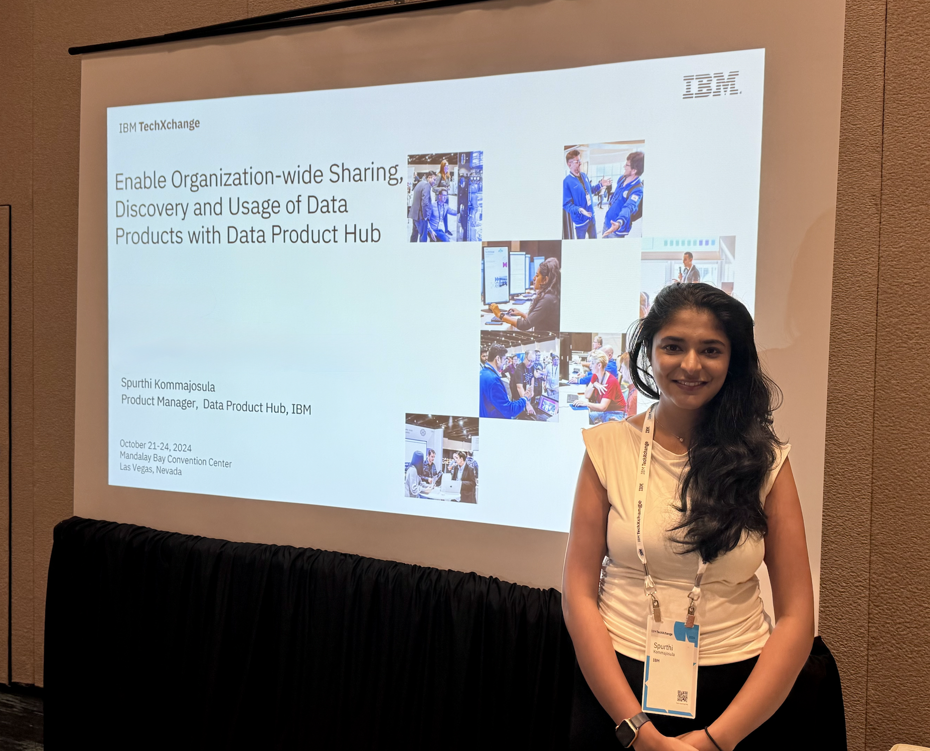 A woman standing in front of a presentation slide at a conference, smiling at the camera. The slide displays information about IBM TechXchange with several small images of people working at computers and attending the event. She is wearing a white top, has a conference badge around her neck and a watch on her wrist.