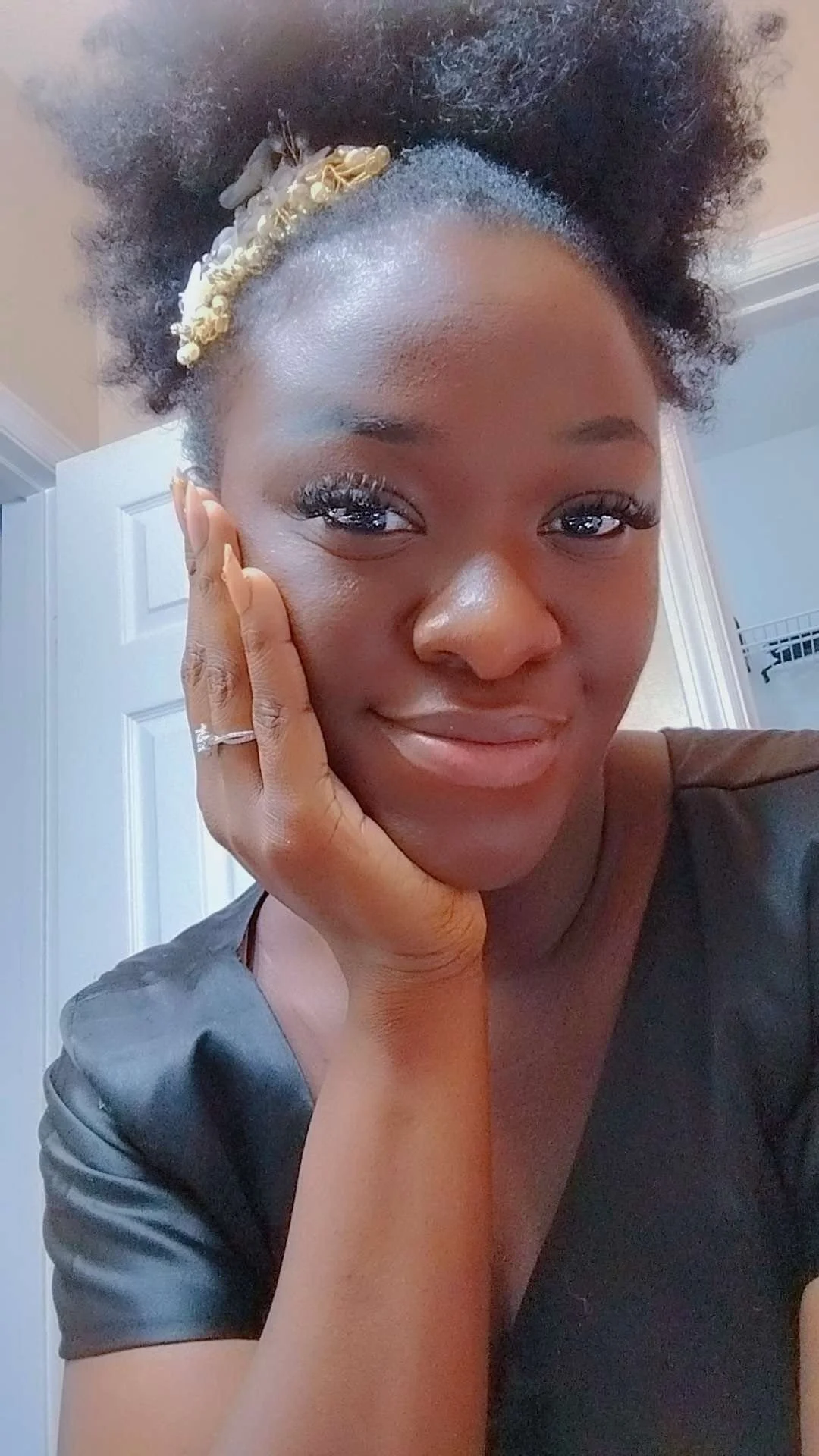 Close-up of a smiling woman with natural hair, adorned with a decorative headband, resting her face on her hand, in an indoor setting.