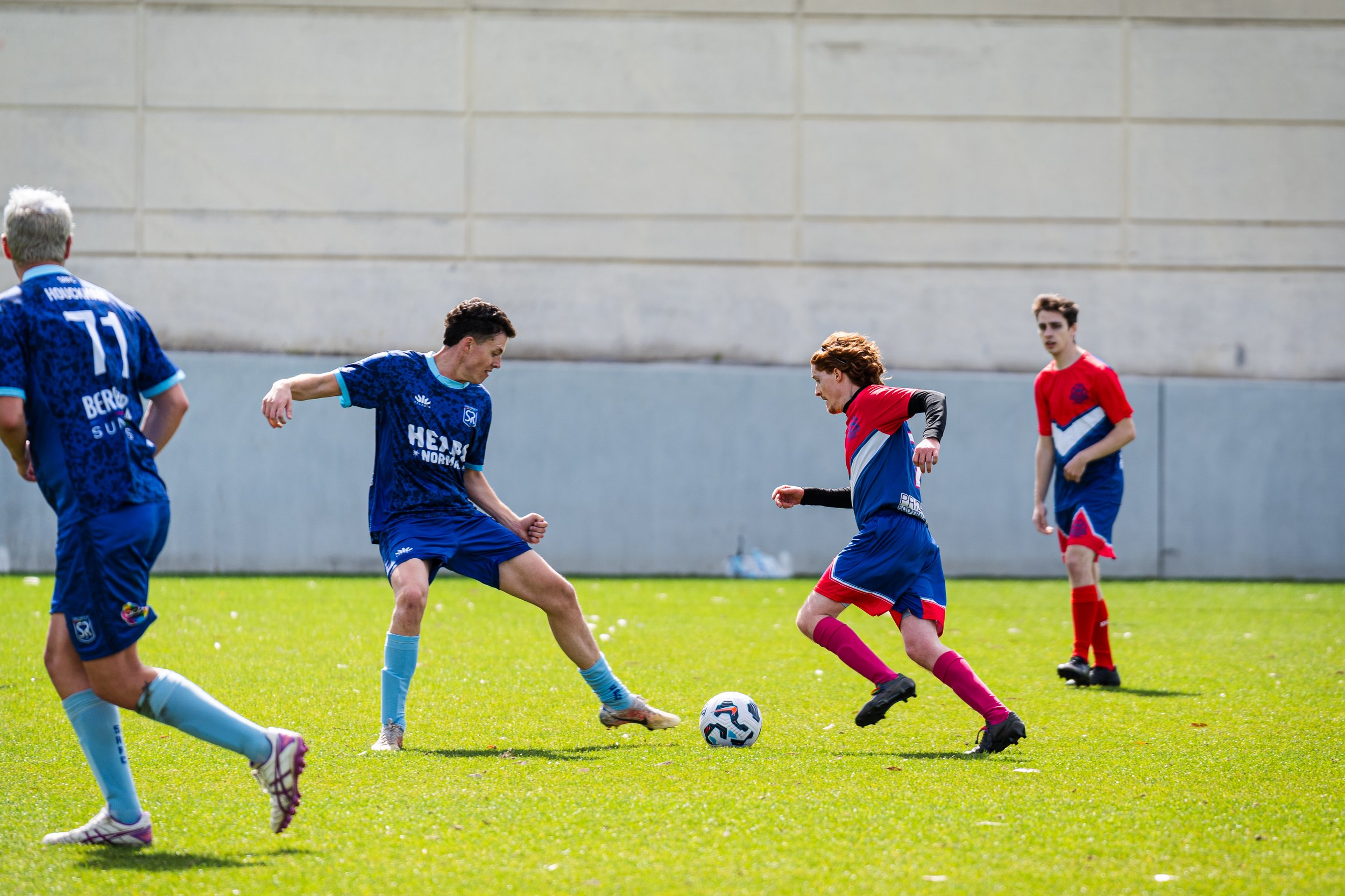 Young soccer players in blue and red uniforms playing on a green field, with one player kicking a soccer ball and others observing.