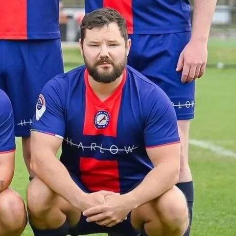 A man with a beard crouching on a soccer field, wearing a blue and red soccer jersey with a logo and the word 'Harlow' on it.