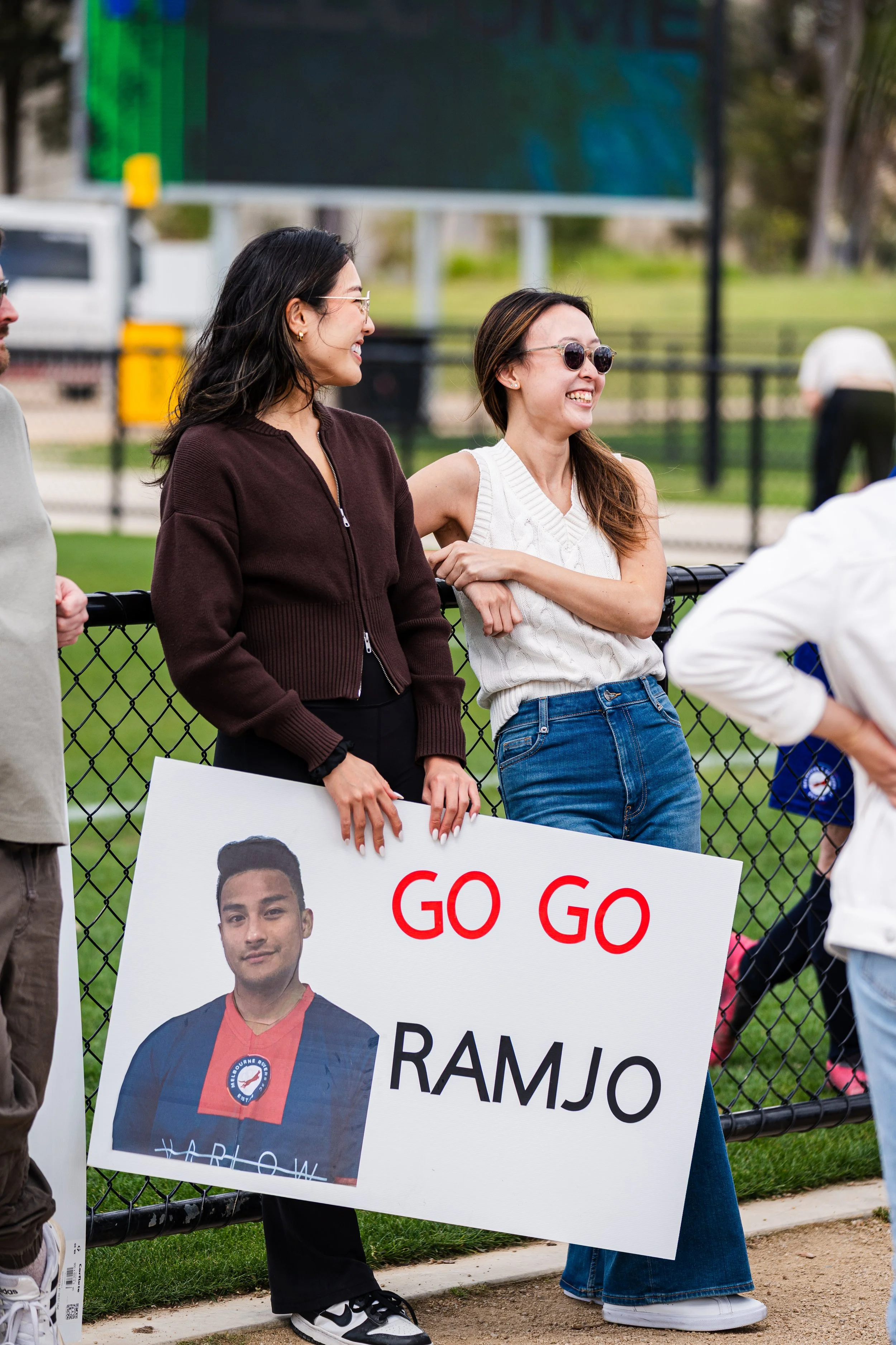 Two women at a sports event, one holding a sign showing a young man in a sports uniform with the message 'GO GO RAMJO'. The women are smiling and wearing casual clothes, standing behind a black fence with a grassy field and a scoreboard in the backgr