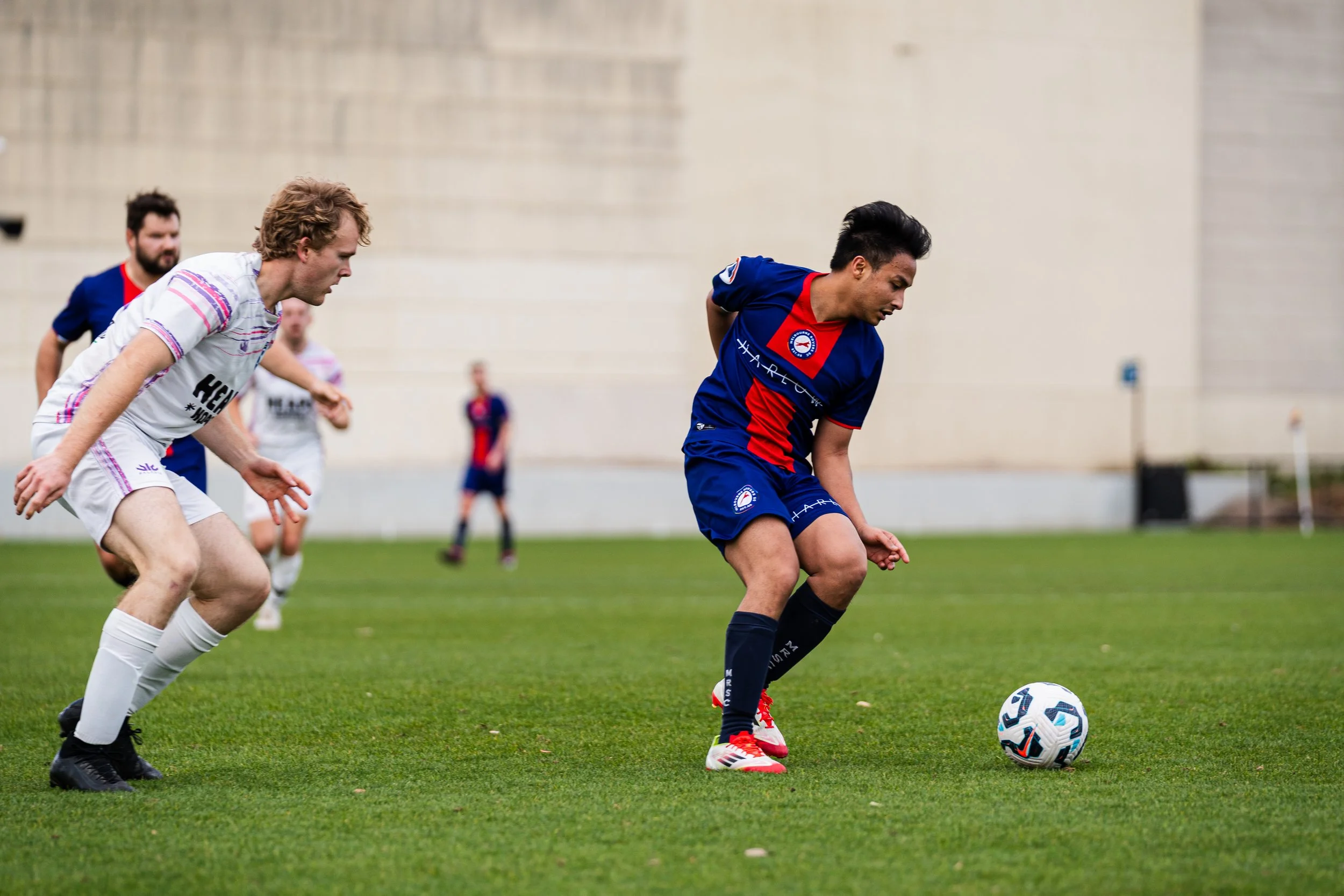 Soccer players competing for the ball on a grass field, with one player in a blue and red uniform and others in white and blue uniforms.