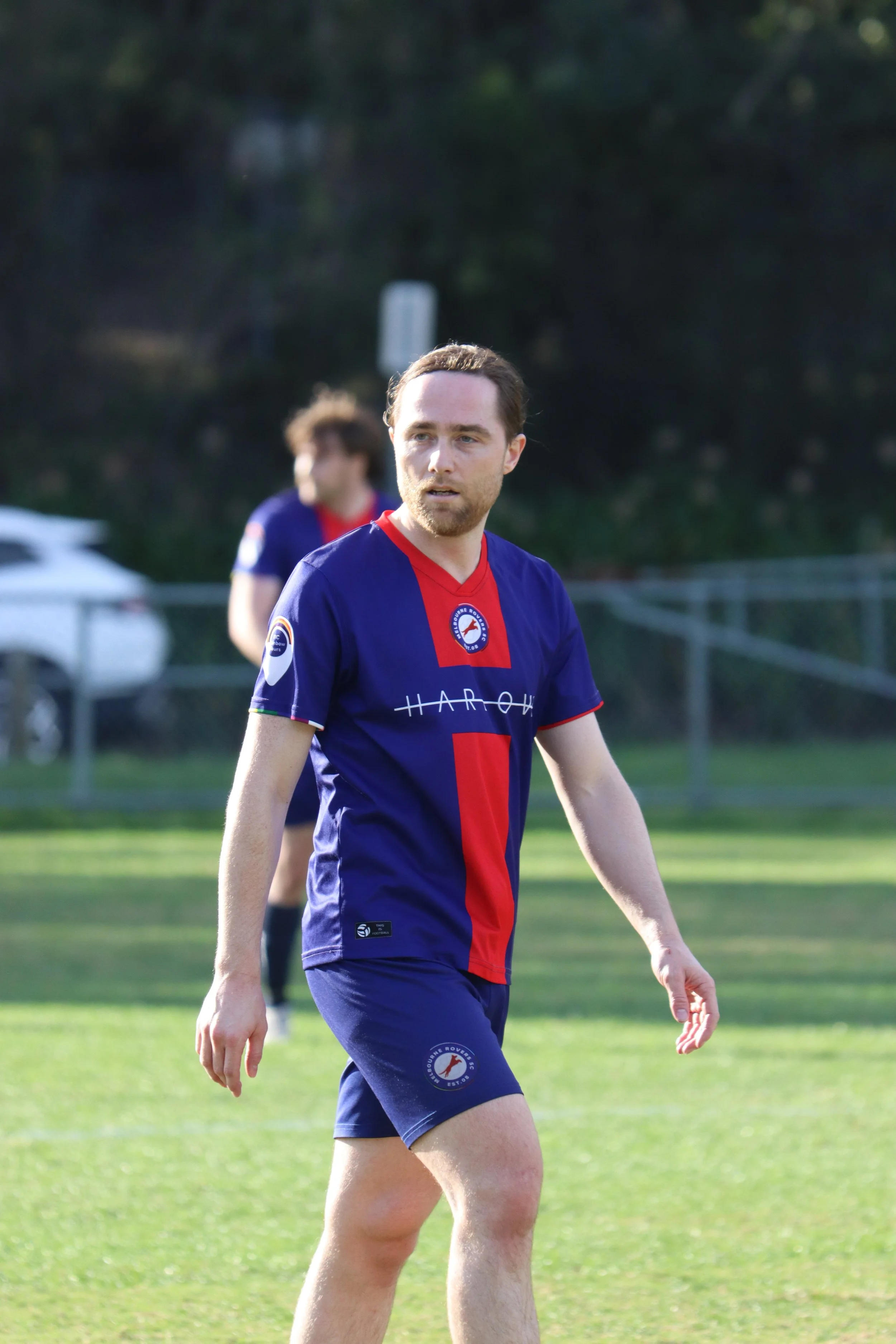 A man in a blue and red soccer jersey walking on a soccer field, with another player blurred in the background.