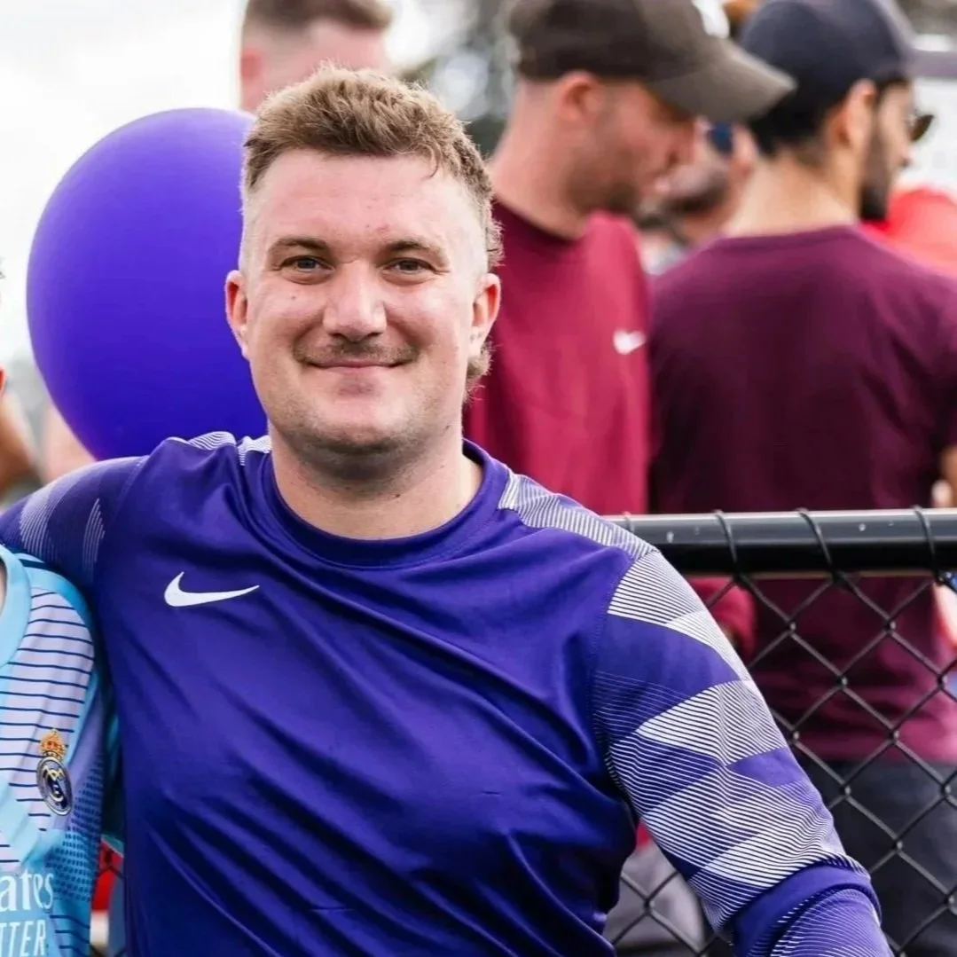 A young man smiling, wearing a blue sports jersey, standing in front of a chain-link fence at an outdoor event, with people in the background.