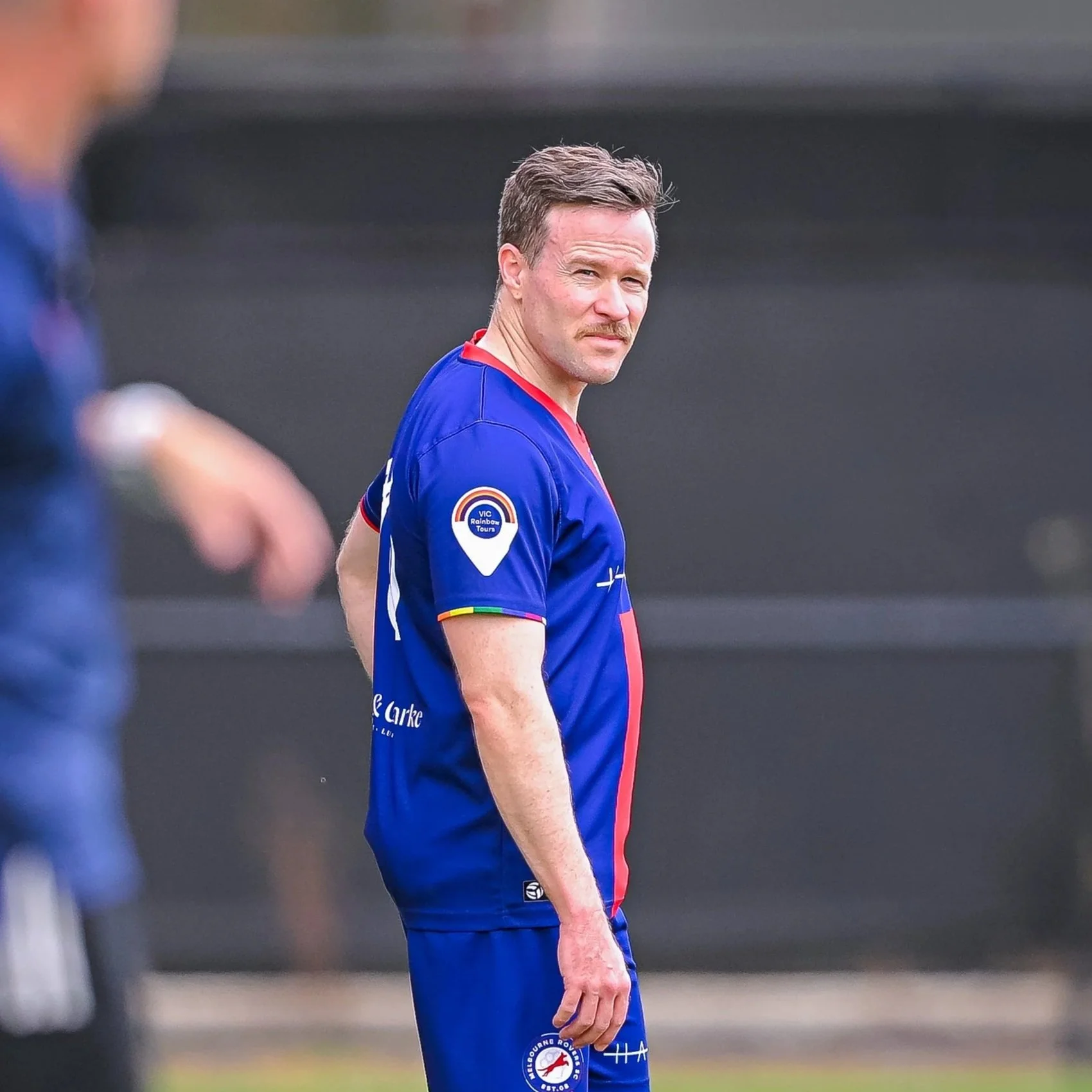 A man with light skin, brown hair, a mustache, wearing a blue sports jersey with red and white accents, standing on a sports field, looking over his shoulder.