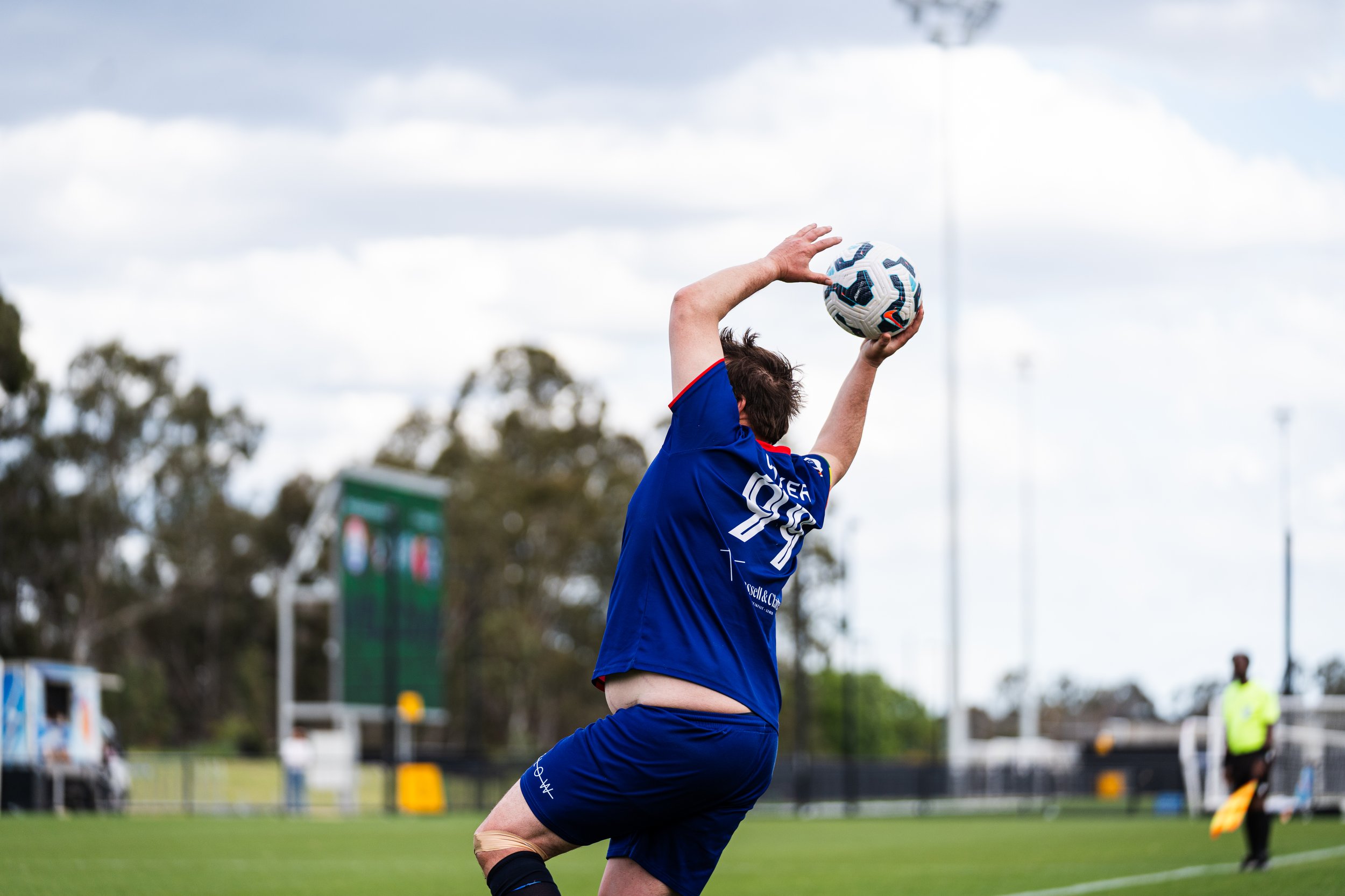 A soccer player in a blue uniform is jumping and preparing to throw a soccer ball during a game on a field with a cloudy sky in the background.