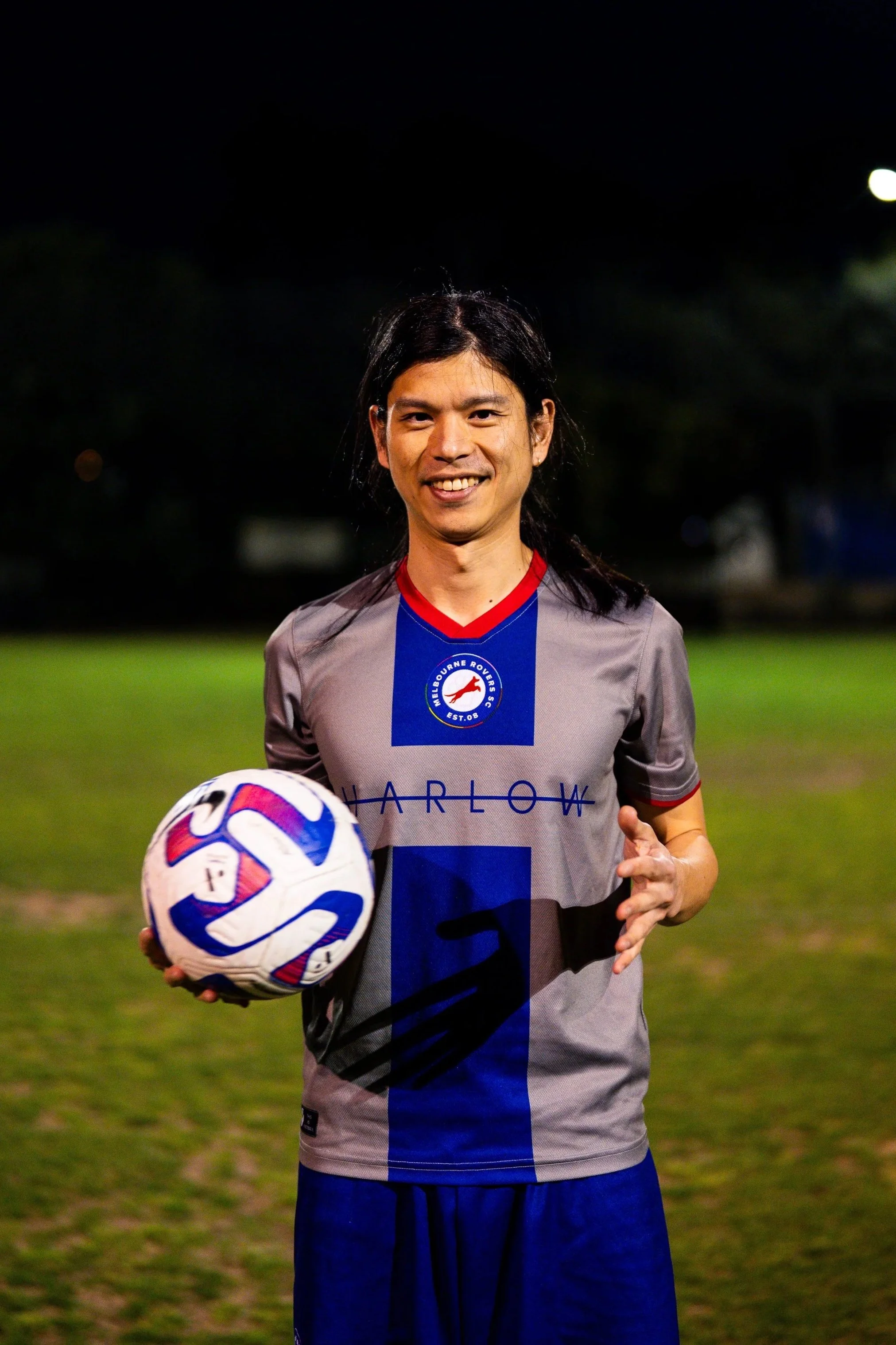 A man in a gray and blue soccer jersey holding a soccer ball on a field at night.