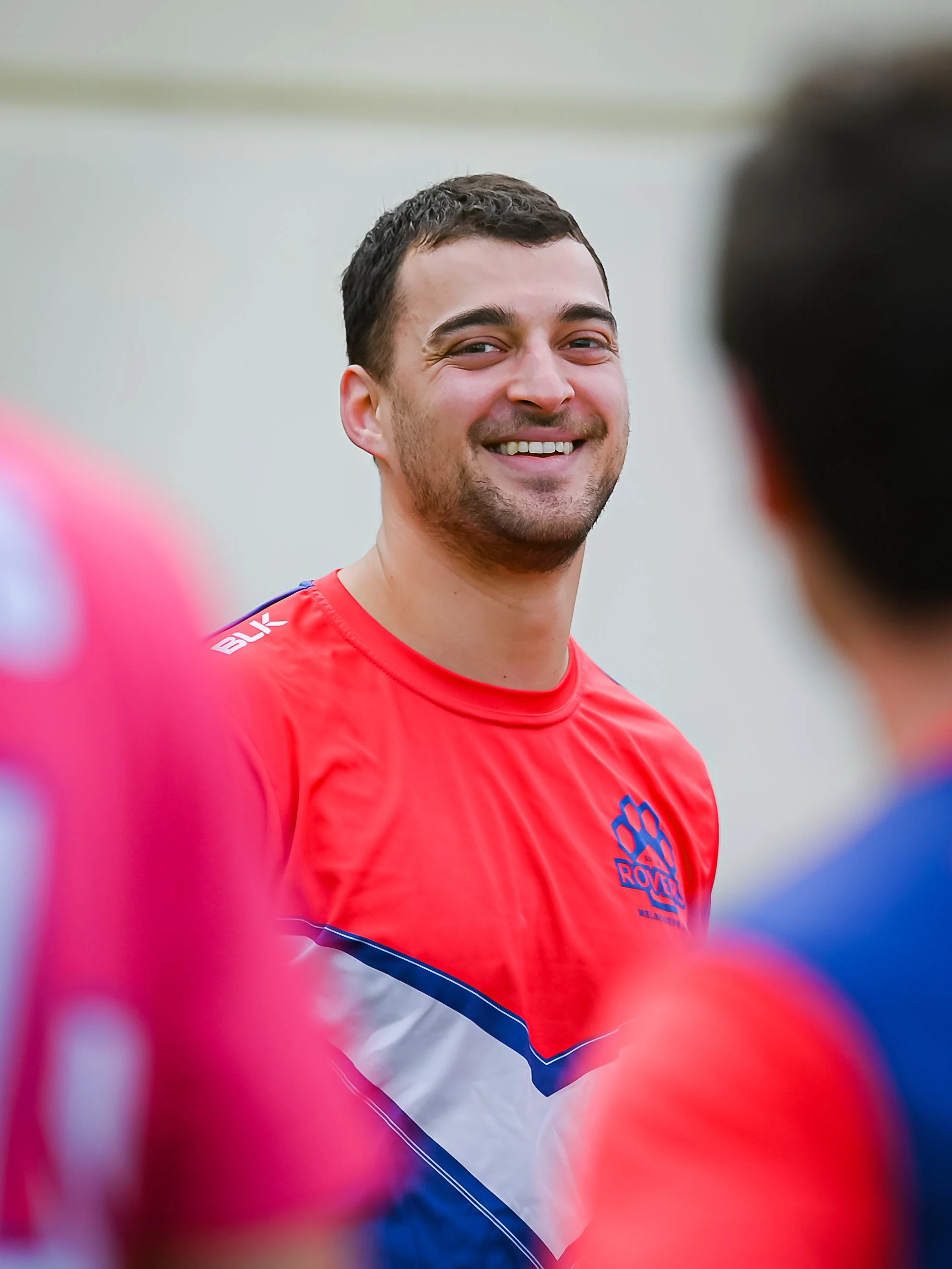 Smiling man in a red sports jersey with a blue and white pattern, standing among other athletes, at a sports event.