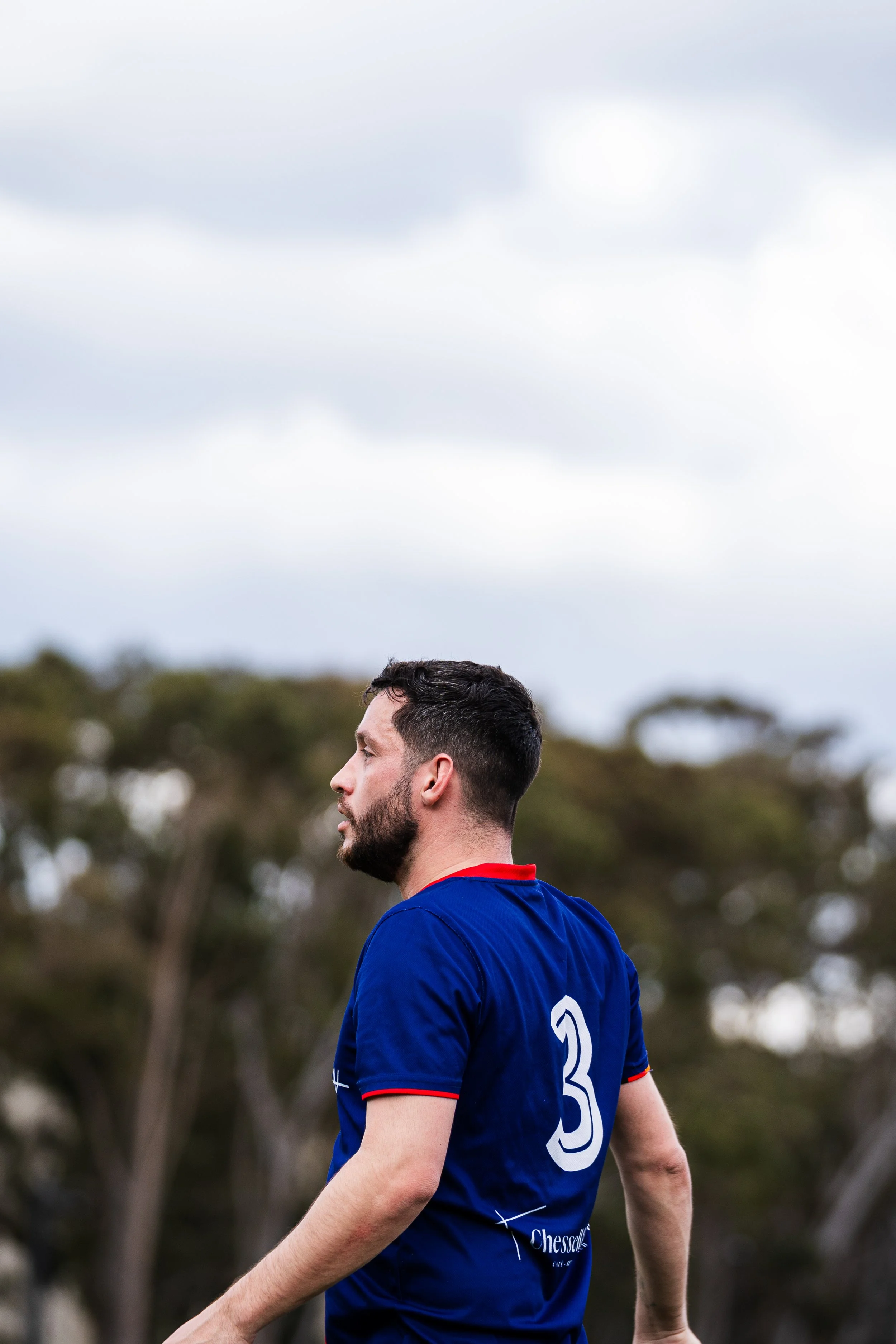Side profile of a man with dark hair and beard wearing a blue sports jersey with the number 3 back, outdoors with trees and cloudy sky in the background.