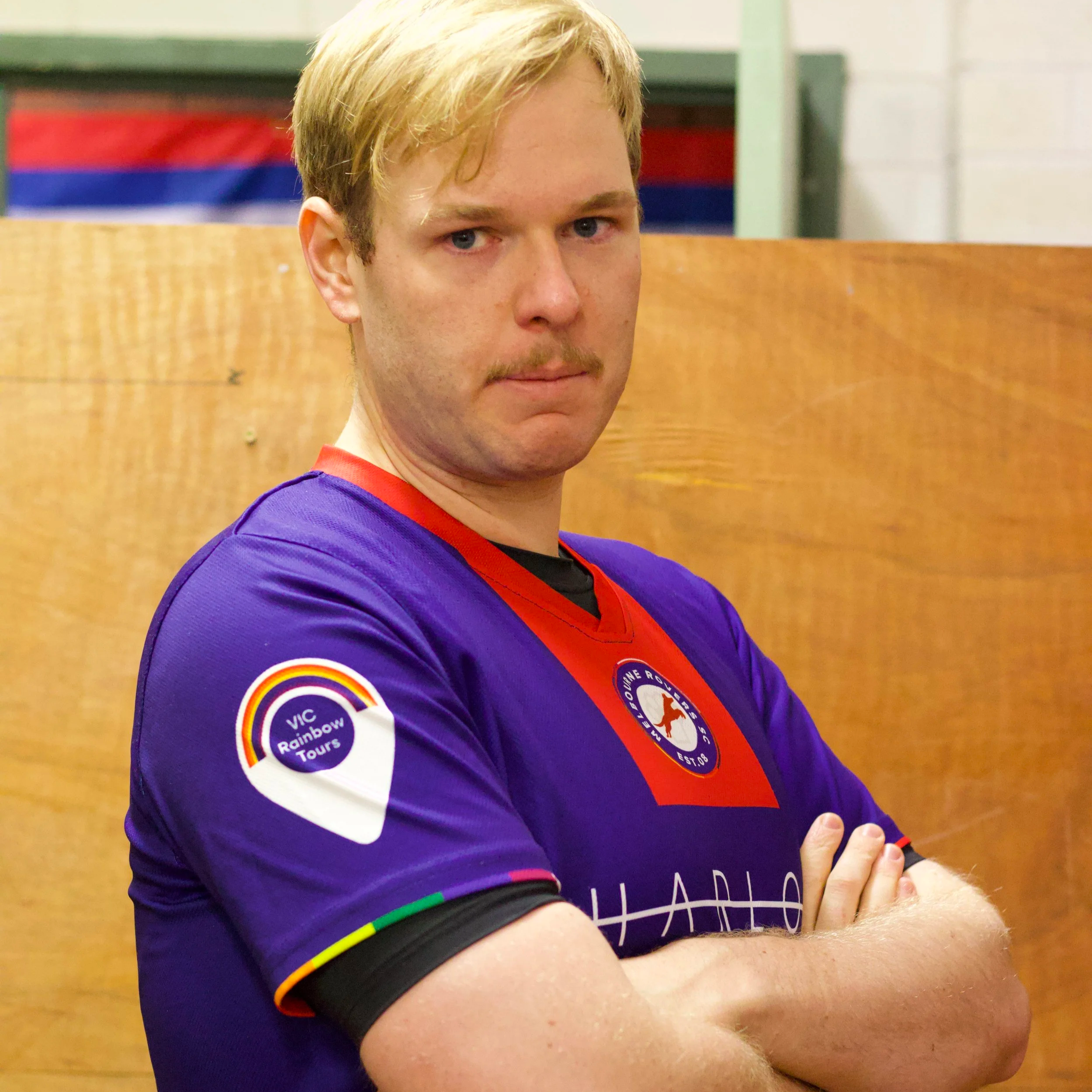 A man with blonde hair and a mustache wearing a purple sports jersey standing with arms crossed in front of a wooden background.