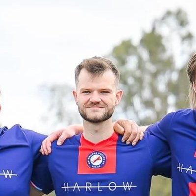 A man wearing a blue and red sports jersey, standing outdoors with his arms around two other people, smiling at the camera.