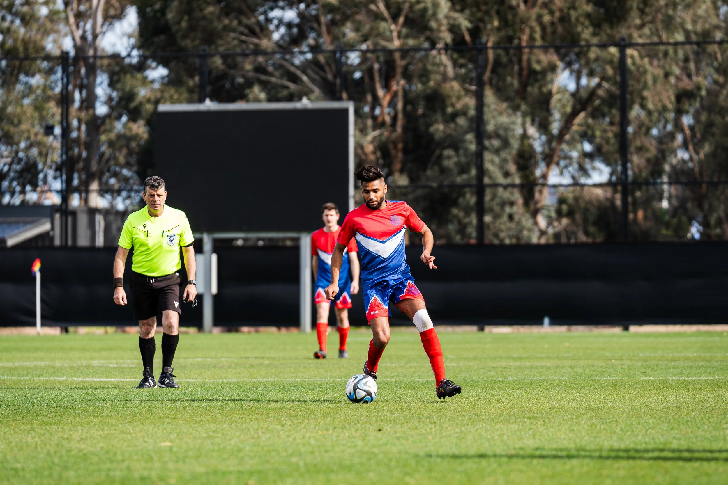 Soccer player in red and blue uniform about to kick a soccer ball on a field, with a referee and another player in the background.