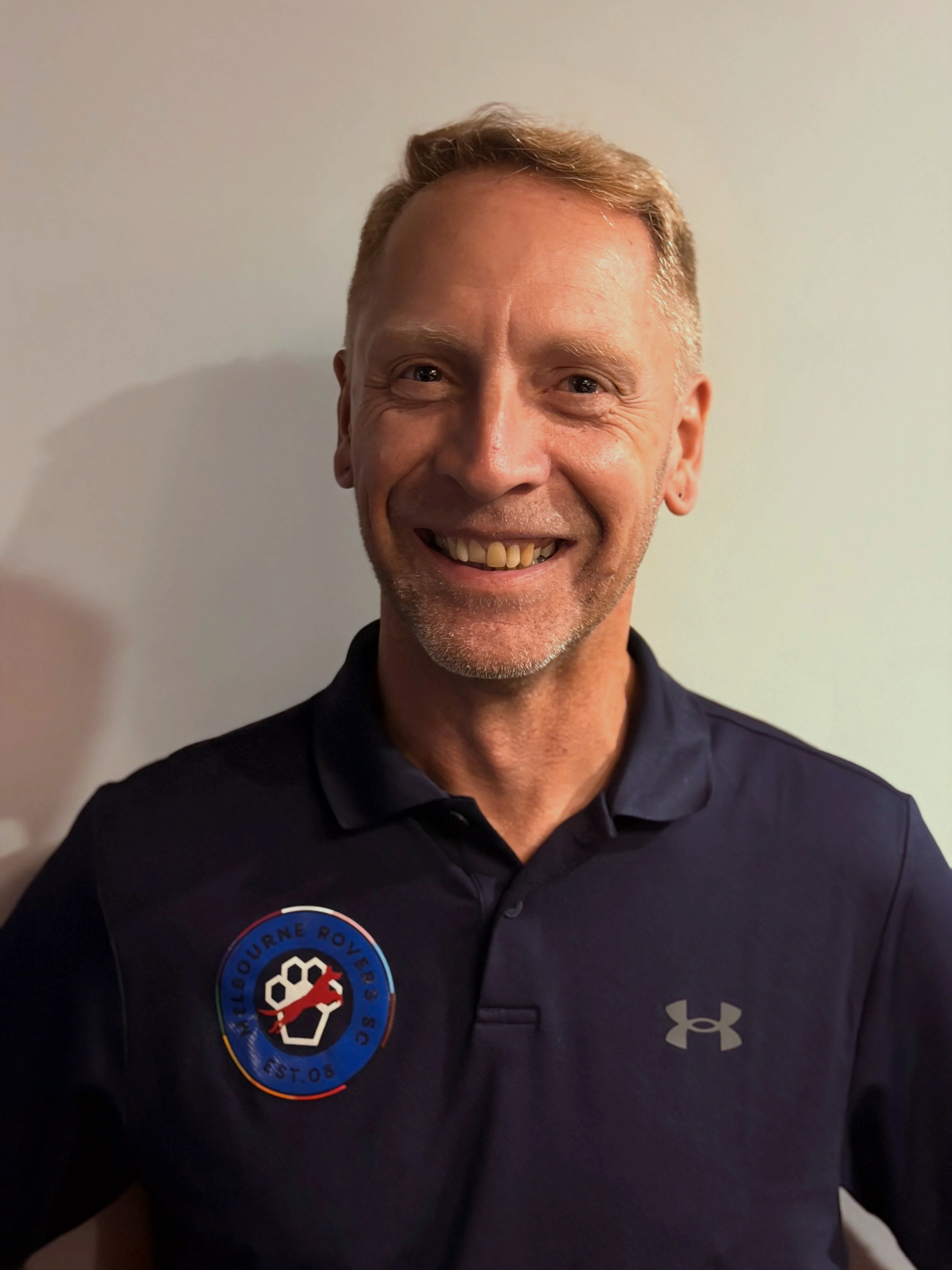 A smiling man with short blonde hair and a trimmed beard, wearing a navy Under Armour polo shirt with a Football Melbourne Rovers SC logo on the chest.