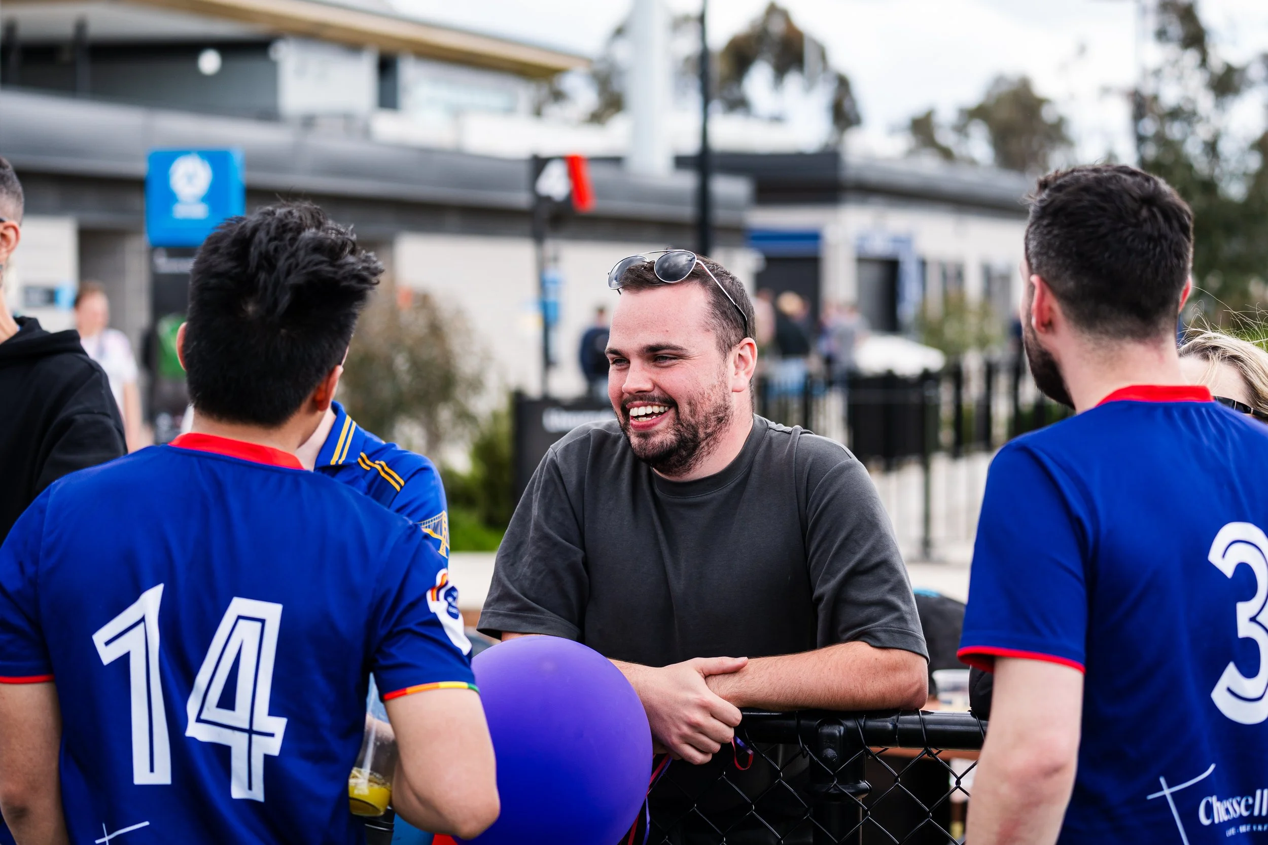 Man in black shirt smiling and talking with three people in blue sports jerseys outside.