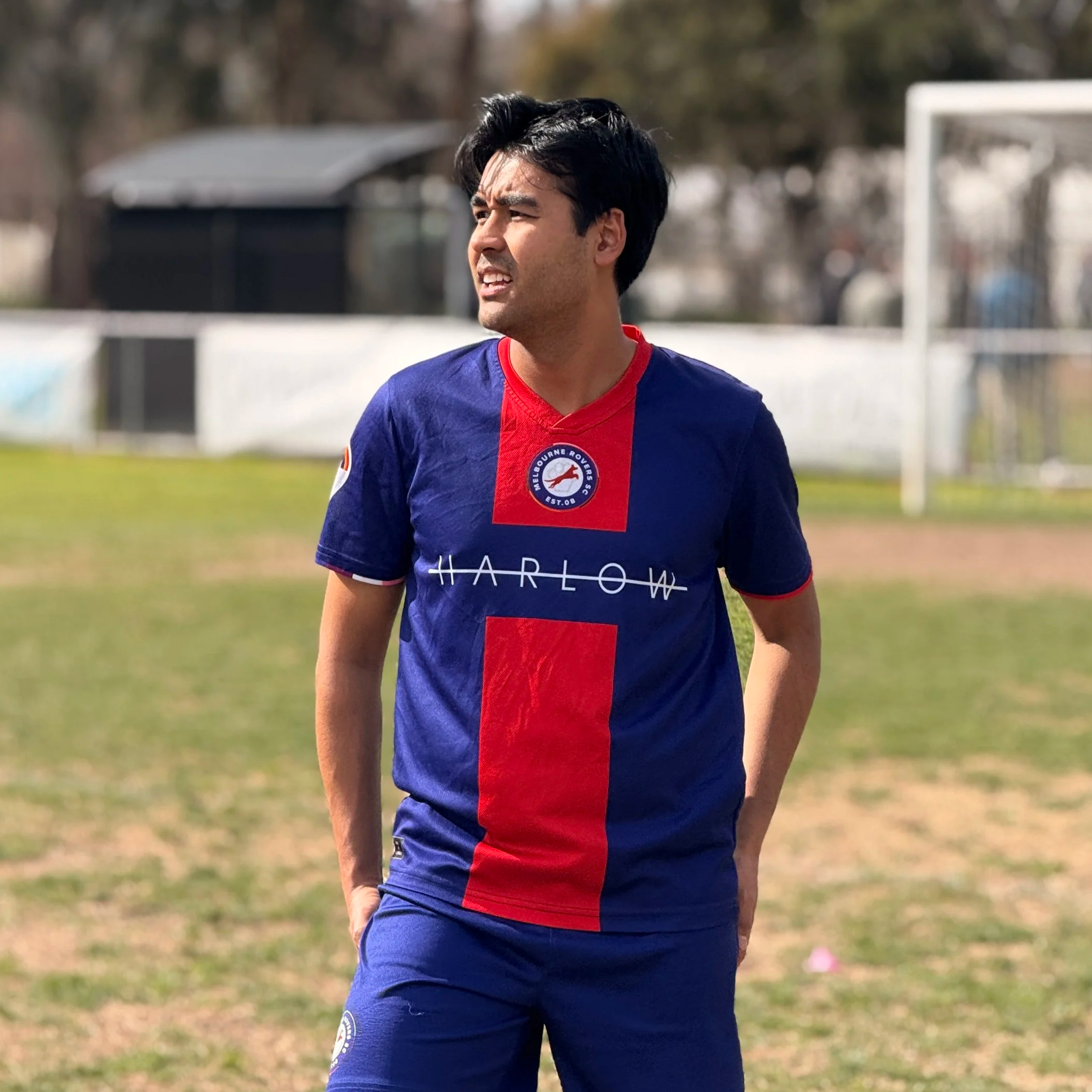A young man with black hair wearing a blue, red, and white sports jersey with the text 'HARLOW' and a patch with a logo, standing on a grassy field with a soccer goal in the background.