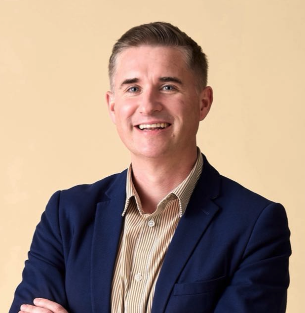 A smiling man in a beige shirt and navy blazer, with arms crossed, against a light beige background.