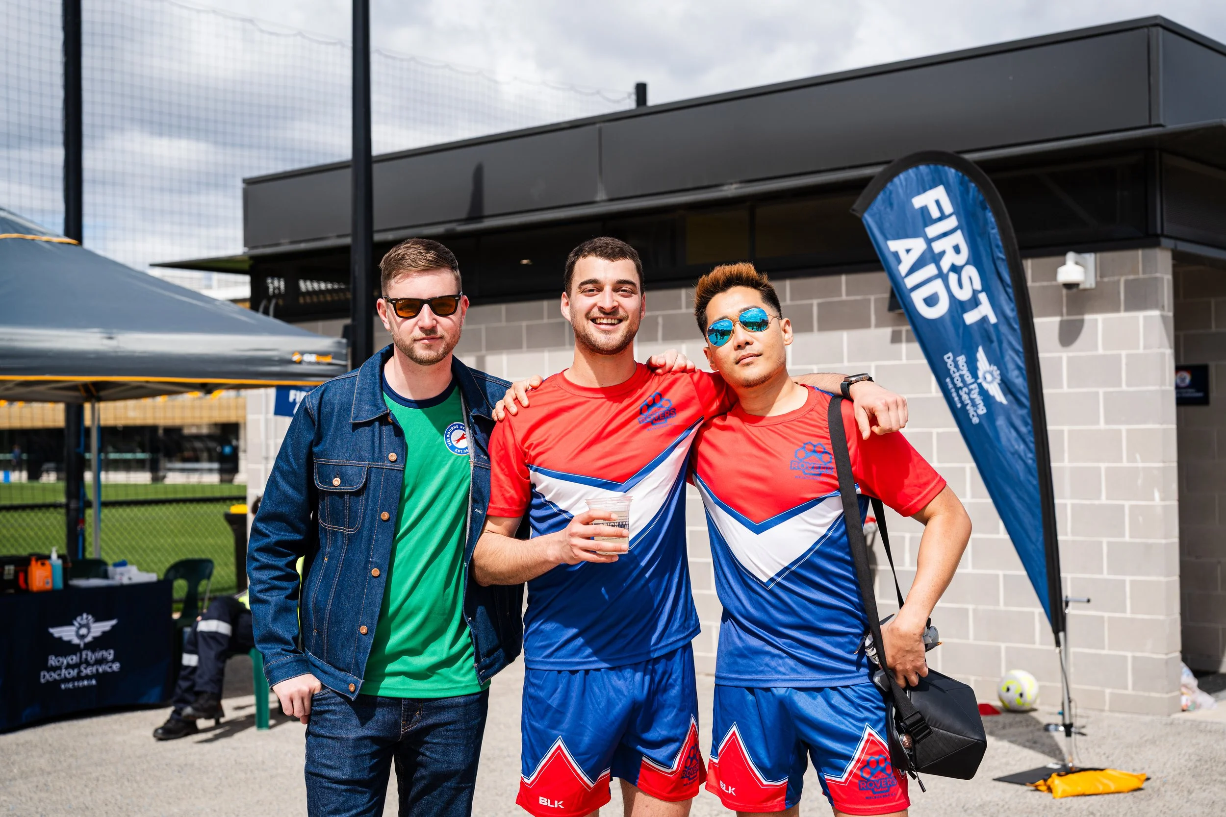 Three young men standing together outdoors, smiling, with one holding a drink. Two of them are wearing red, blue, and white sports jerseys, and one is wearing a green t-shirt with a denim jacket. They are at an event with a tent and a flag that reads
