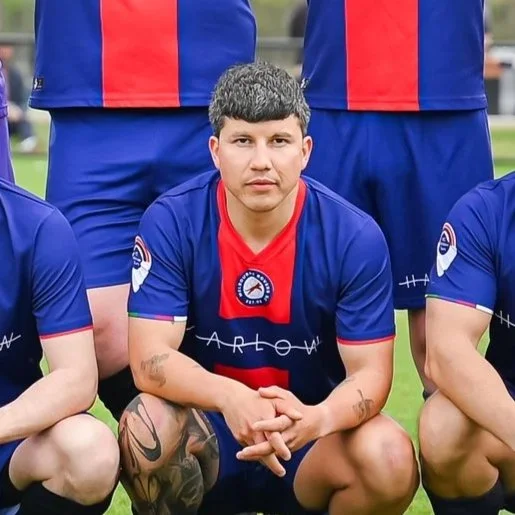 A young man with tattoos on his arms, crouching on a soccer field, wearing a blue and red soccer jersey, posing with teammates.