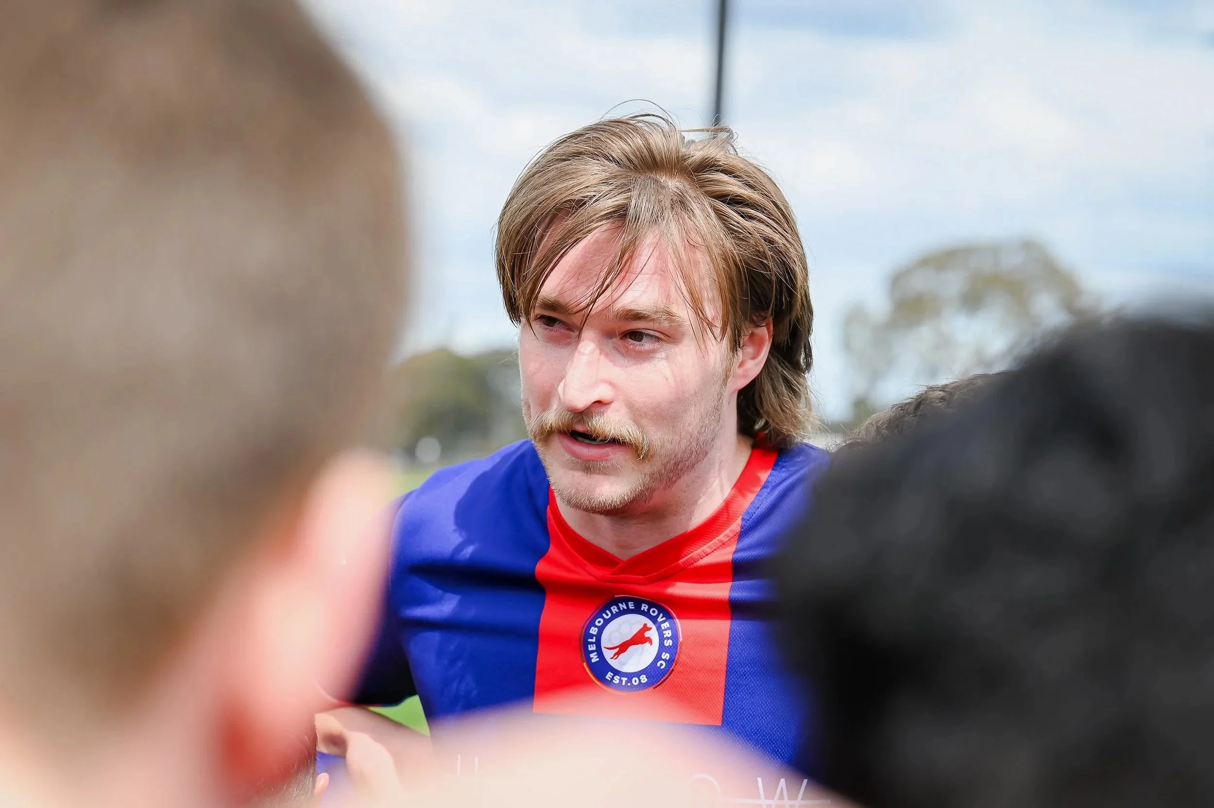 A man with shoulder-length brown hair and a mustache, wearing a blue and red sports jersey with the Melbourne Rovers FC logo, speaking during a sports event.