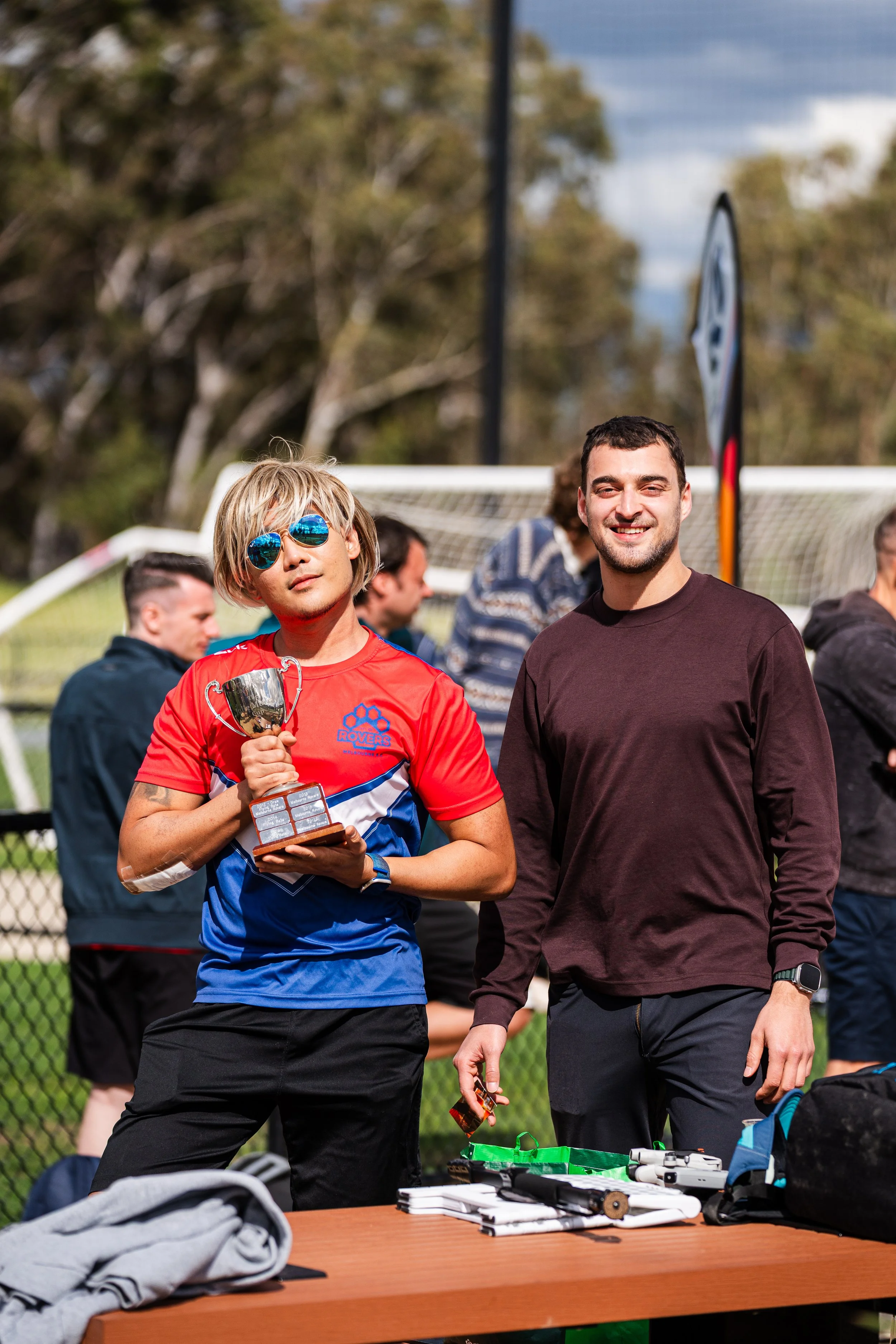 A person holding a trophy standing next to another person at an outdoor event near a soccer field, with people and a goal in the background.