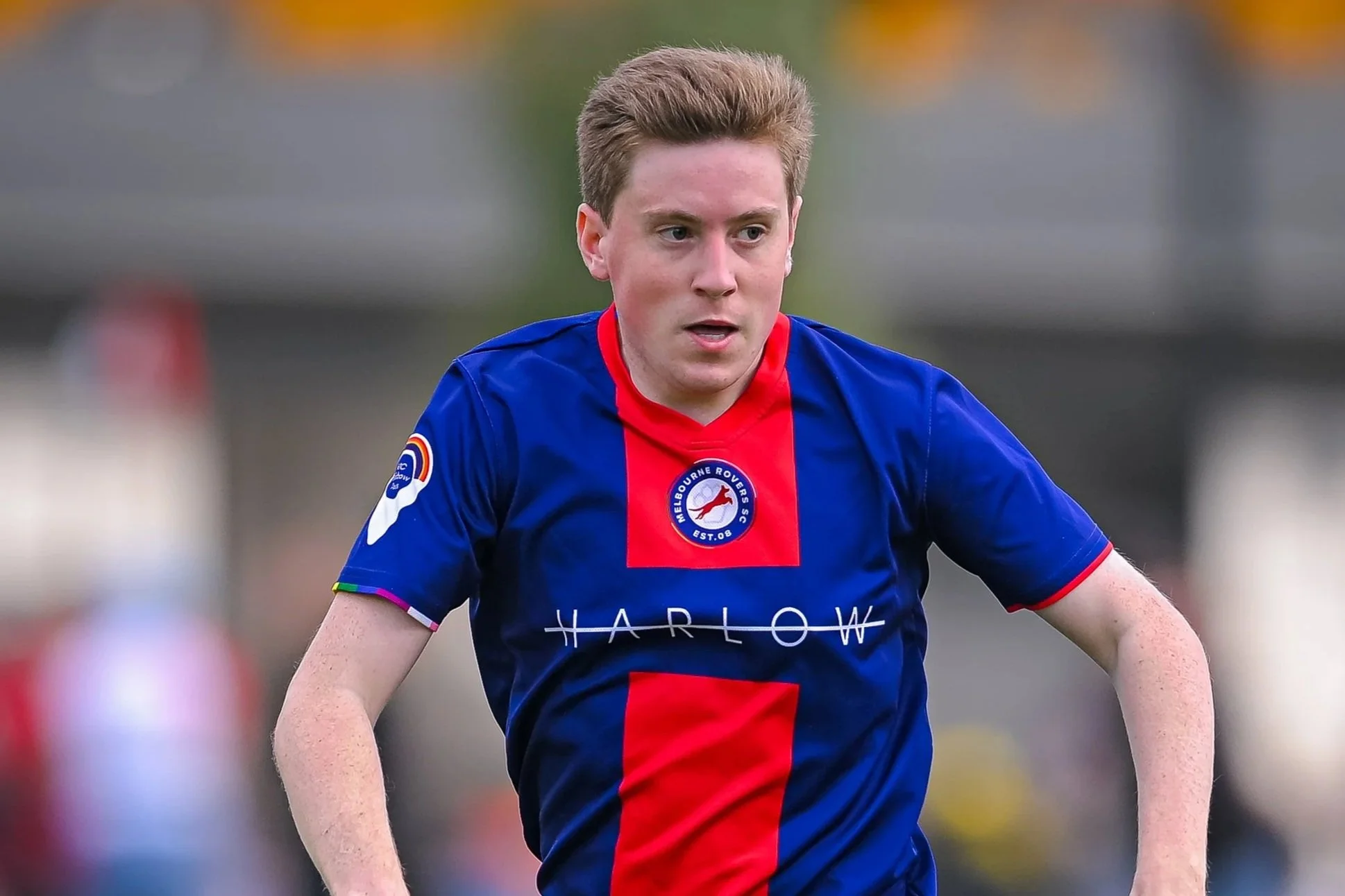 A young man with light brown hair, wearing a blue sports jersey with red accents, is playing soccer outside. The jersey has a Melbourne Rovers badge and a sponsor logo that says Harlow.