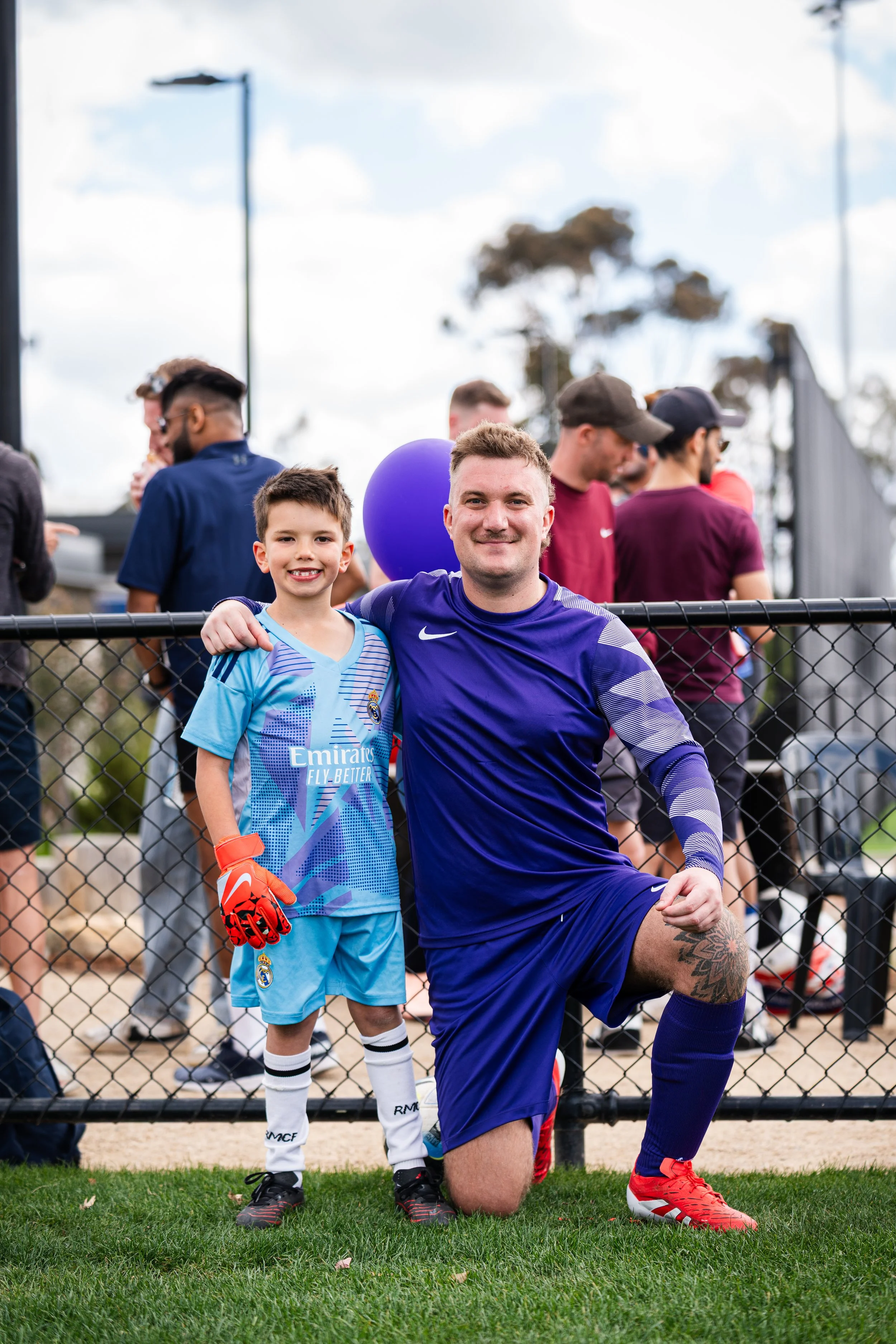 A young boy and an adult man kneeling on a grass field, smiling at the camera during a soccer event. The boy wears a blue soccer jersey, white socks, and soccer cleats, holding orange goalkeeper gloves. The man is dressed in a blue soccer uniform wit