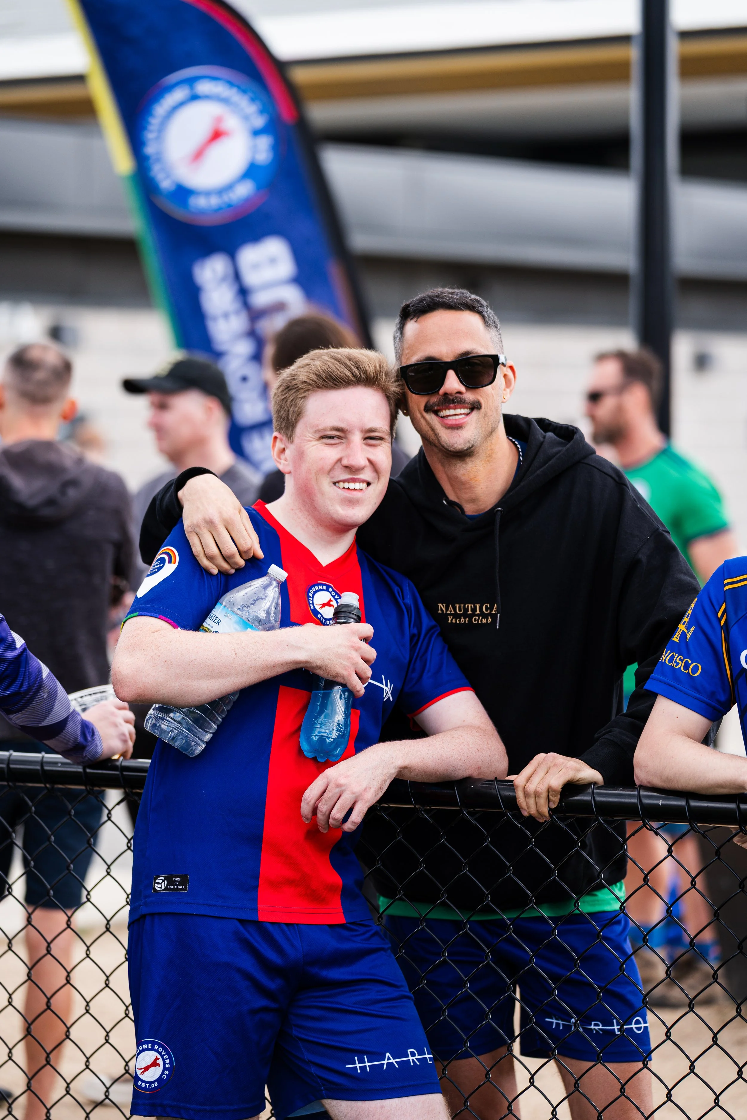 Two men smiling and posing together at a sports event, one wearing a blue and red sports jersey and the other in a black hoodie, with a crowd and a banner in the background.