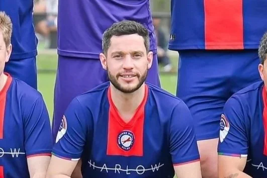 A group of soccer players in blue and red jerseys, with one player kneeling in front, smiling at the camera.