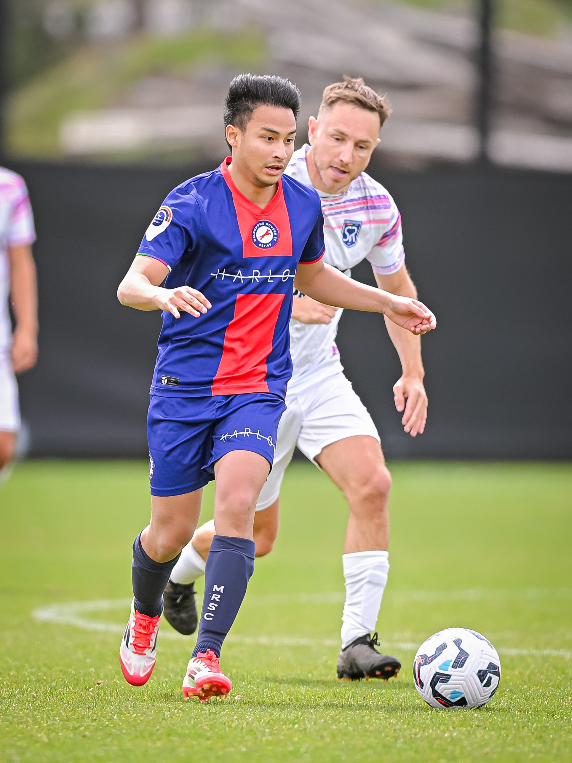 Two soccer players competing for the ball on the field. The player in the foreground is wearing a blue and red jersey with matching shorts and socks, while the player behind is dressed in a white and purple kit.