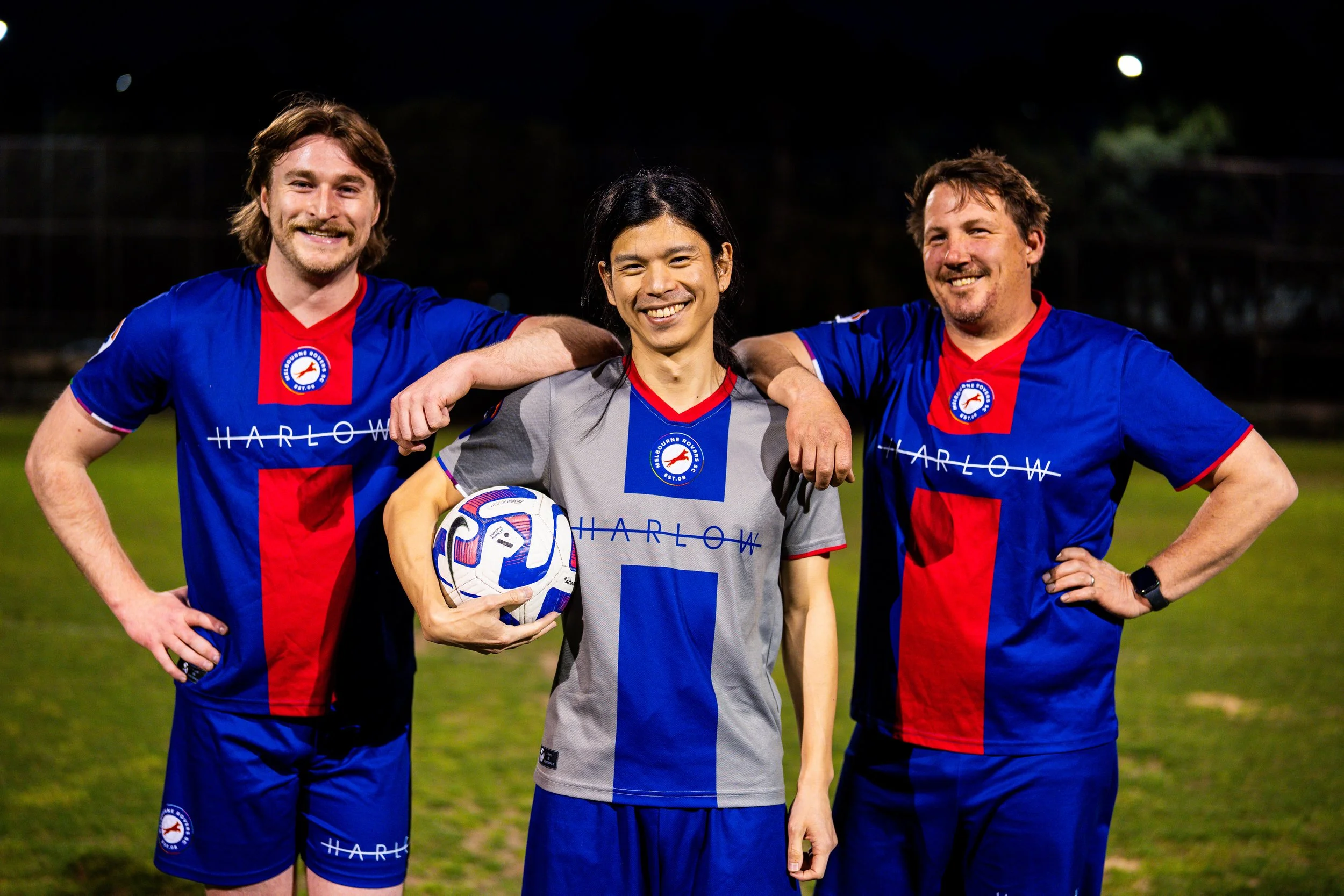 Three men in soccer jerseys standing on a field at night, smiling, with their arms around each other's shoulders. The man in the middle is holding a soccer ball.
