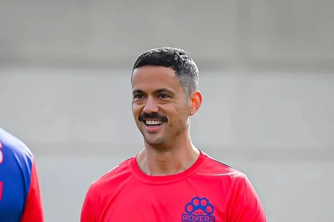 A smiling man with short dark hair and a mustache wearing a red sports jersey with a blue emblem and the word "Rovers".