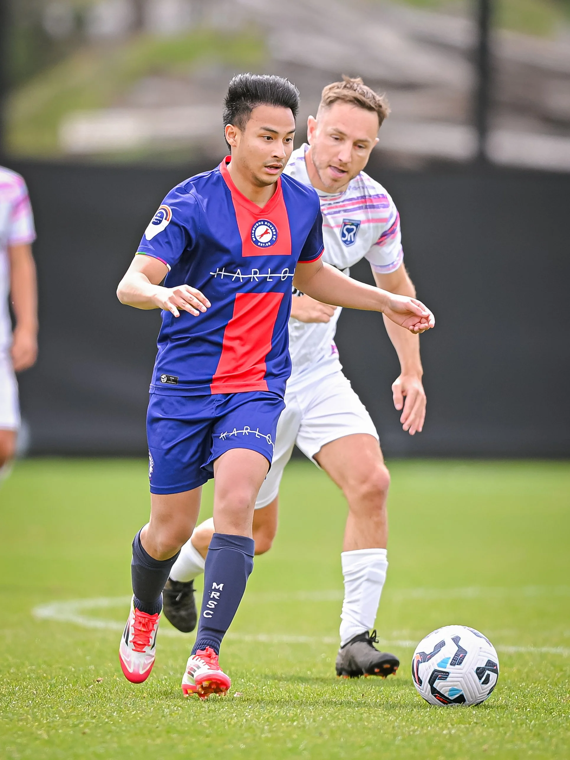 Two soccer players compete for the ball on a grassy field, one wearing a blue and red uniform and the other in white with colorful stripes.