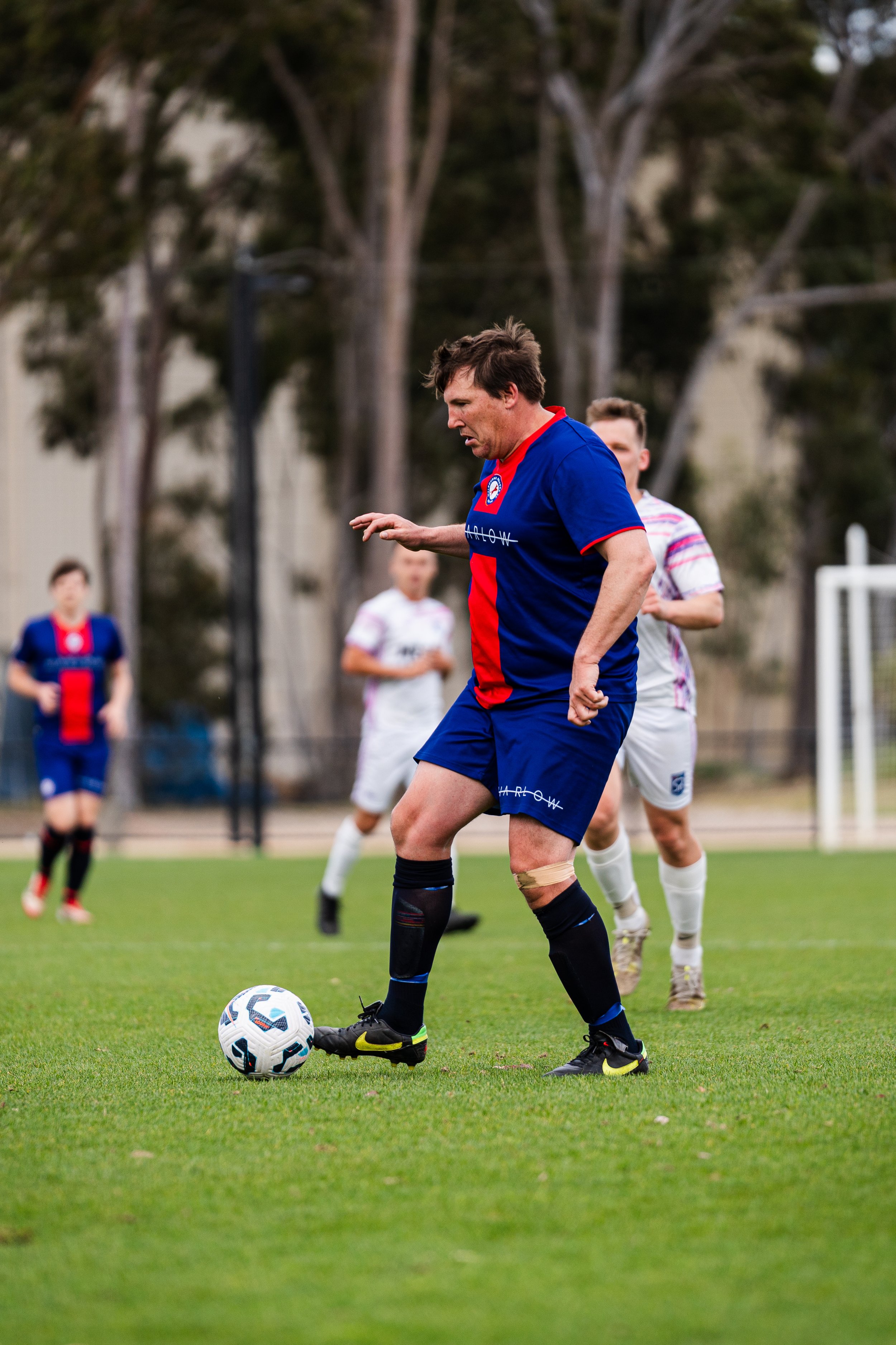 A man in a blue and red soccer jersey and shorts with black knee-high socks standing on a soccer field, about to kick a soccer ball, with other players and trees in the background.