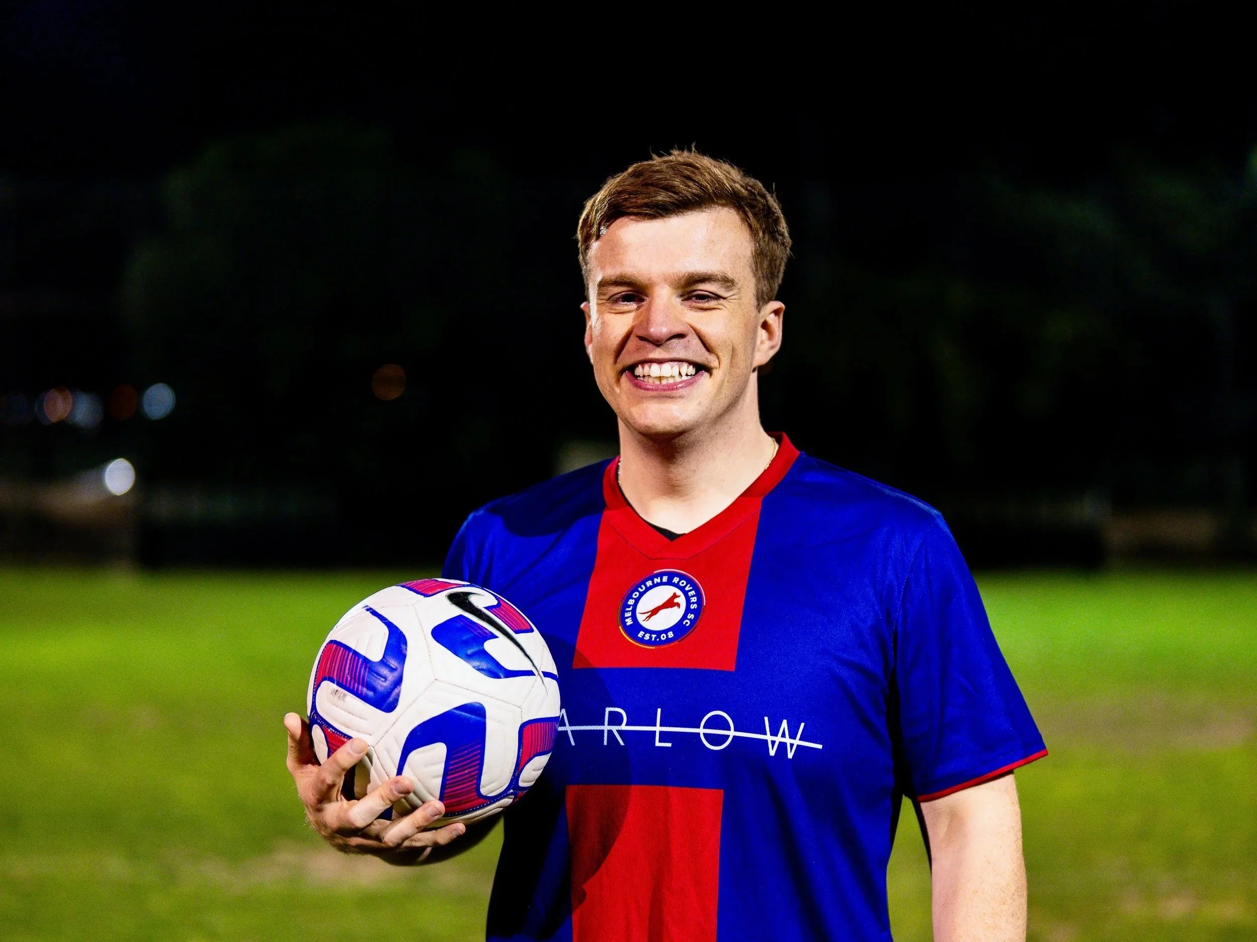 A young man in a blue and red soccer jersey holding a soccer ball on a soccer field at night, smiling.