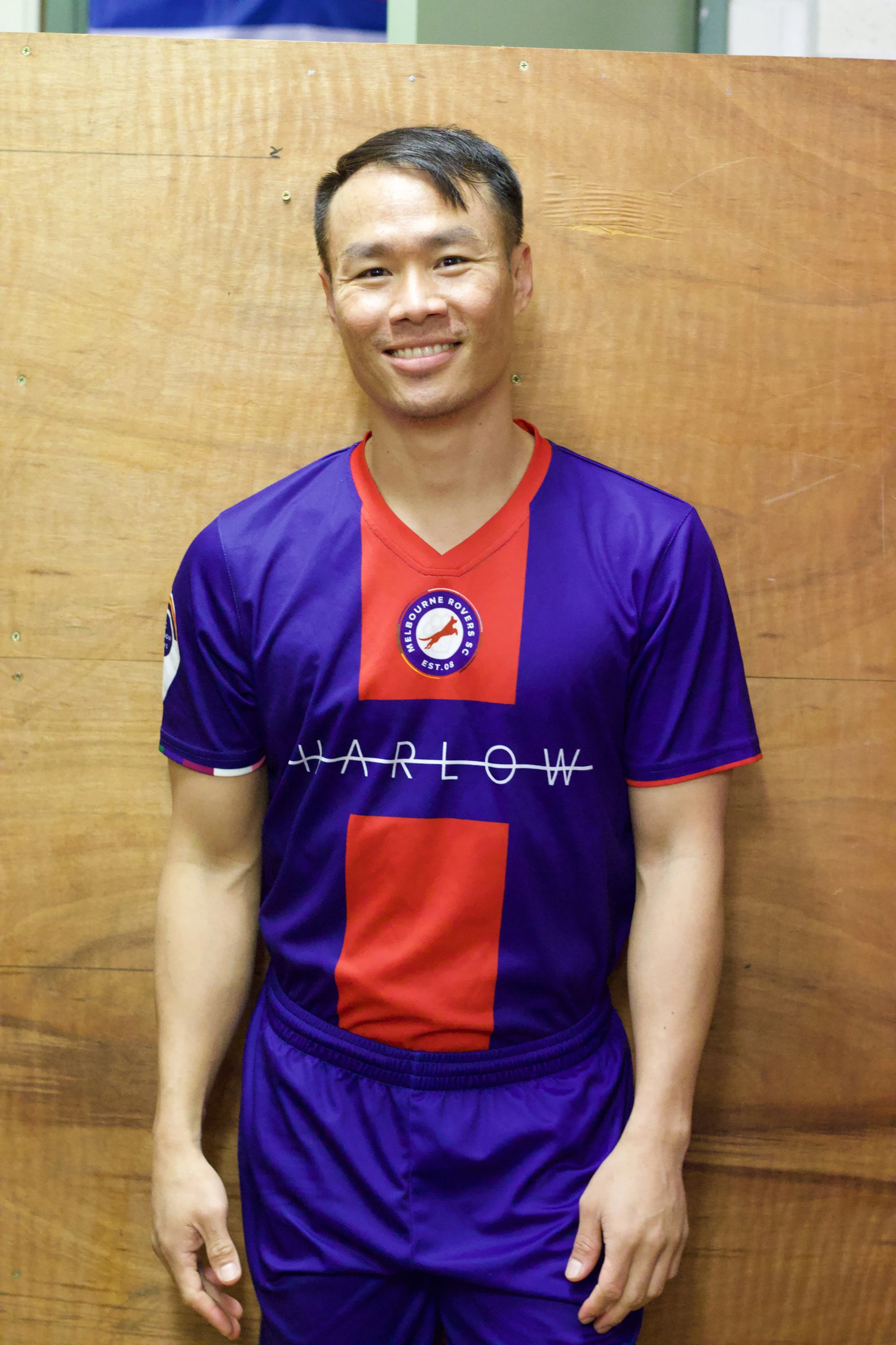 A man standing in front of a wooden wall, wearing a purple and red sports jersey with a logo of Melbourne Rovers on the chest, smiling at the camera.