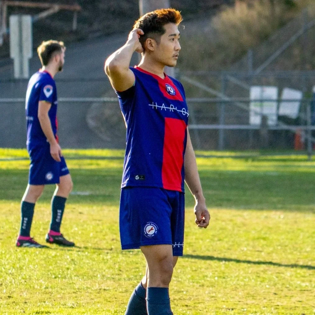 Two soccer players standing on a grassy field during sunset, wearing blue and red uniforms. The player in the foreground has short dark hair and is adjusting his hair, while the player in the background is looking to the left.