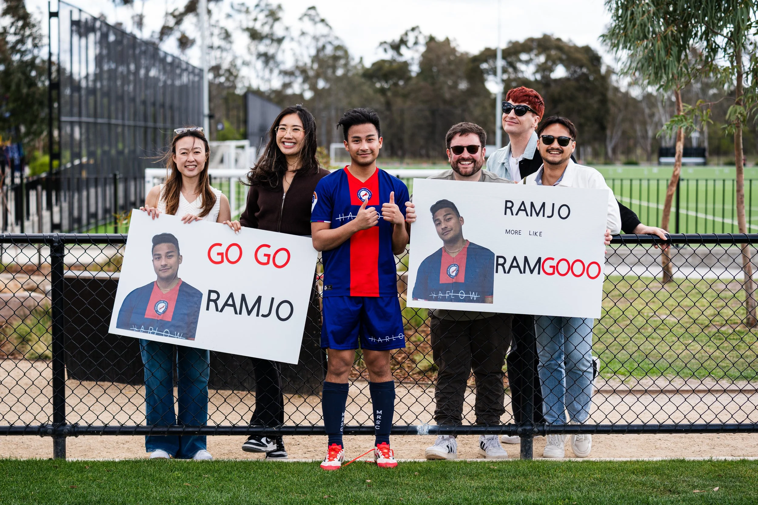 Group of six people standing behind a black metal fence, holding signs to support a person named Ramjo, with a grassy sports field and trees in the background.