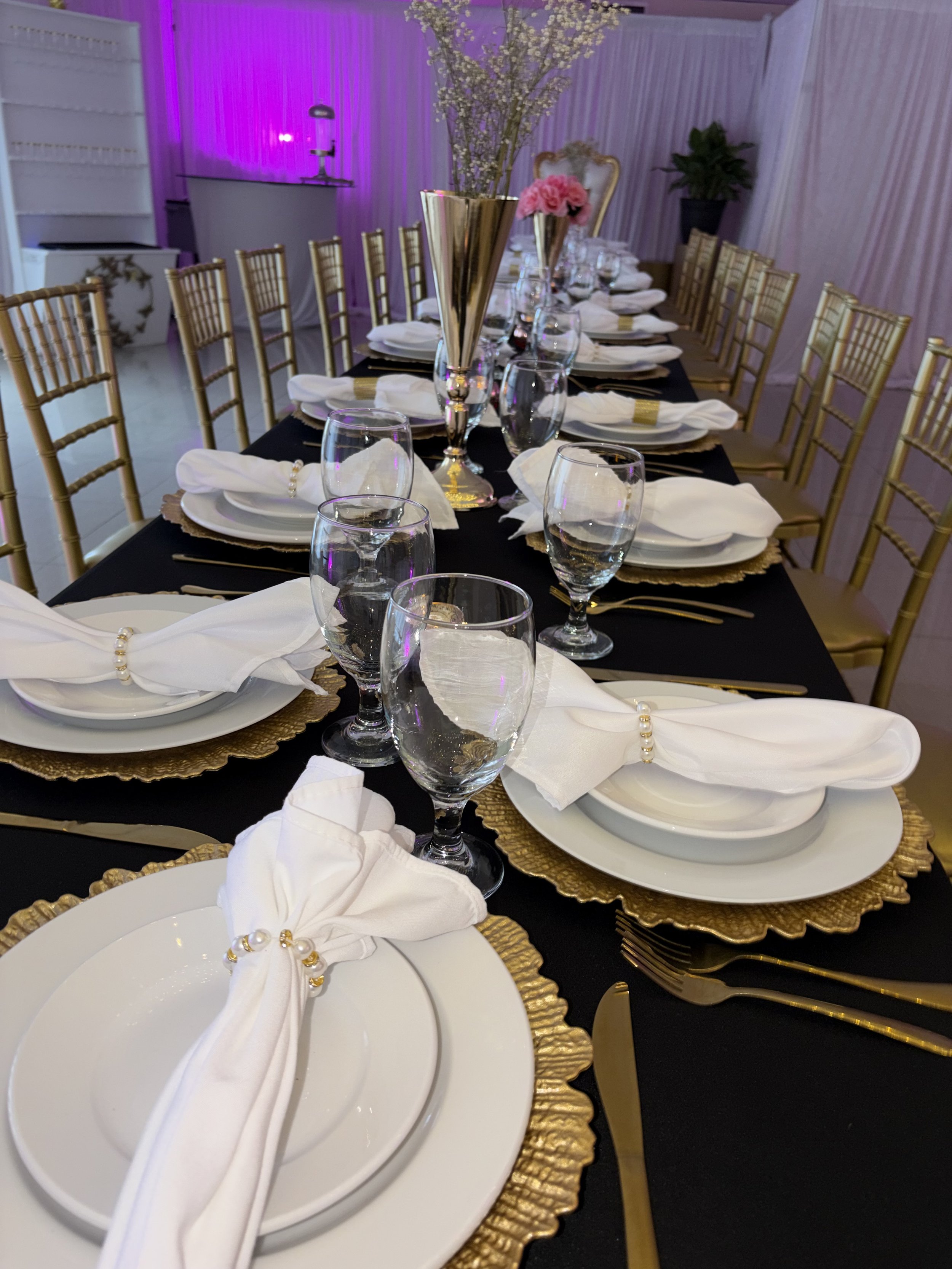 A formal dining table set with white plates, gold chargers, white napkins with pearl napkin rings, multiple wine glasses, and a centerpiece with tall gold vases filled with pink and white flowers. The table has a black tablecloth, and gold chairs are arranged around it. The background features pink and purple lighting, decorative plants, and a pink curtain.