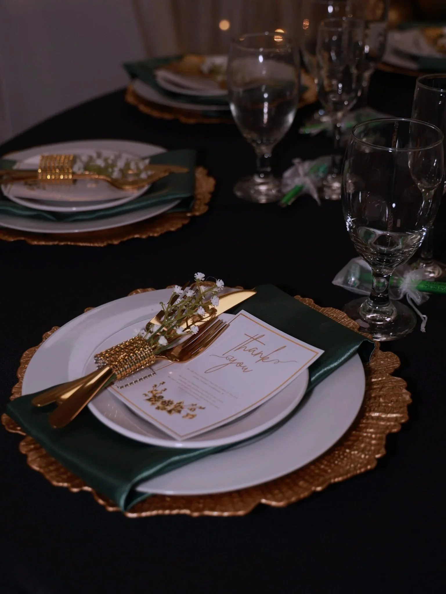 A formal dinner table setting with white plates, gold cutlery, a green napkin, a thank you card, and multiple glasses on a dark tablecloth.