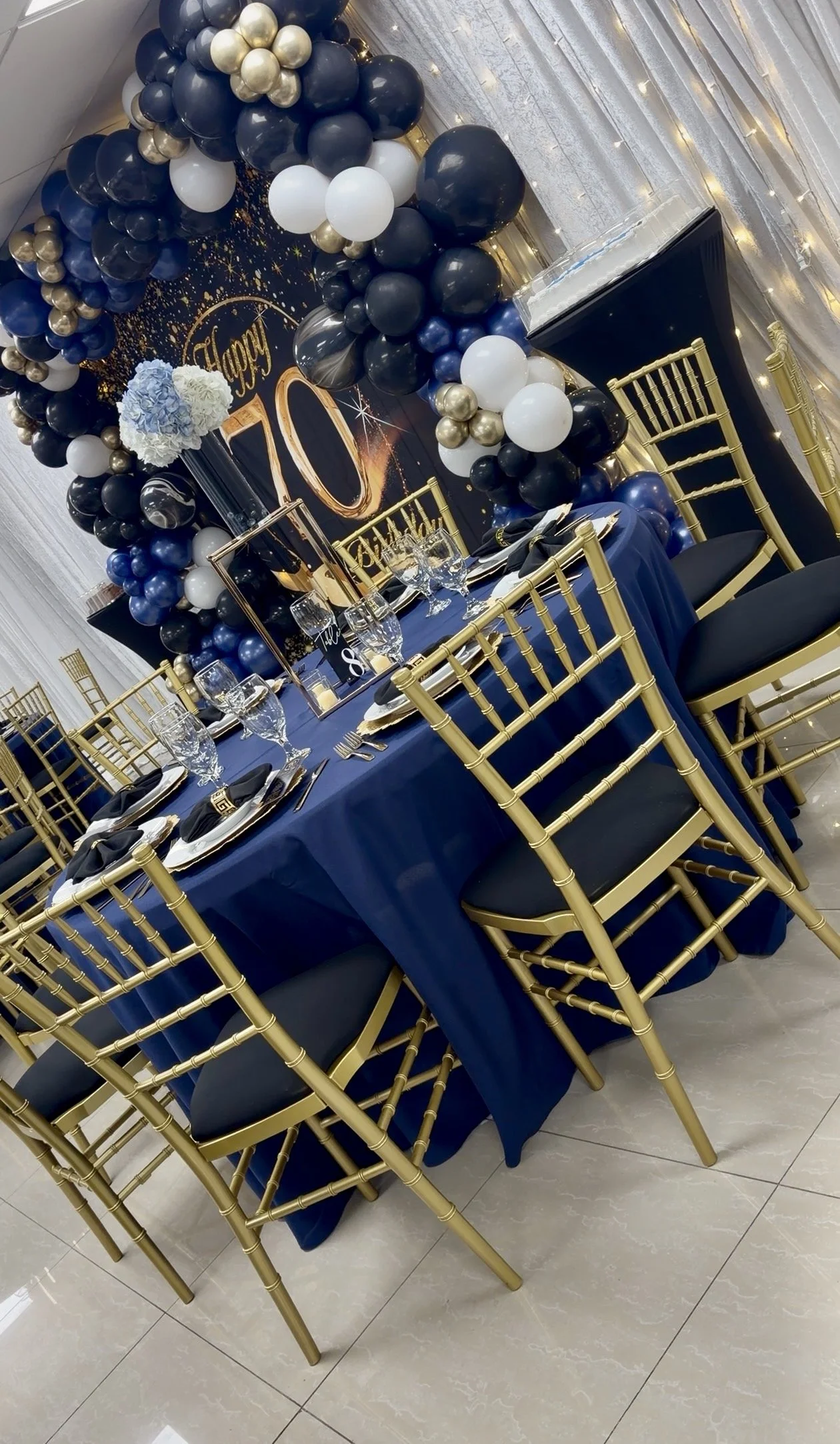 An elegant banquet table decor with a navy blue tablecloth, gold chairs, and a backdrop adorned with navy blue, white, black, and gold balloons, along with fairy lights and a large white floral arrangement.