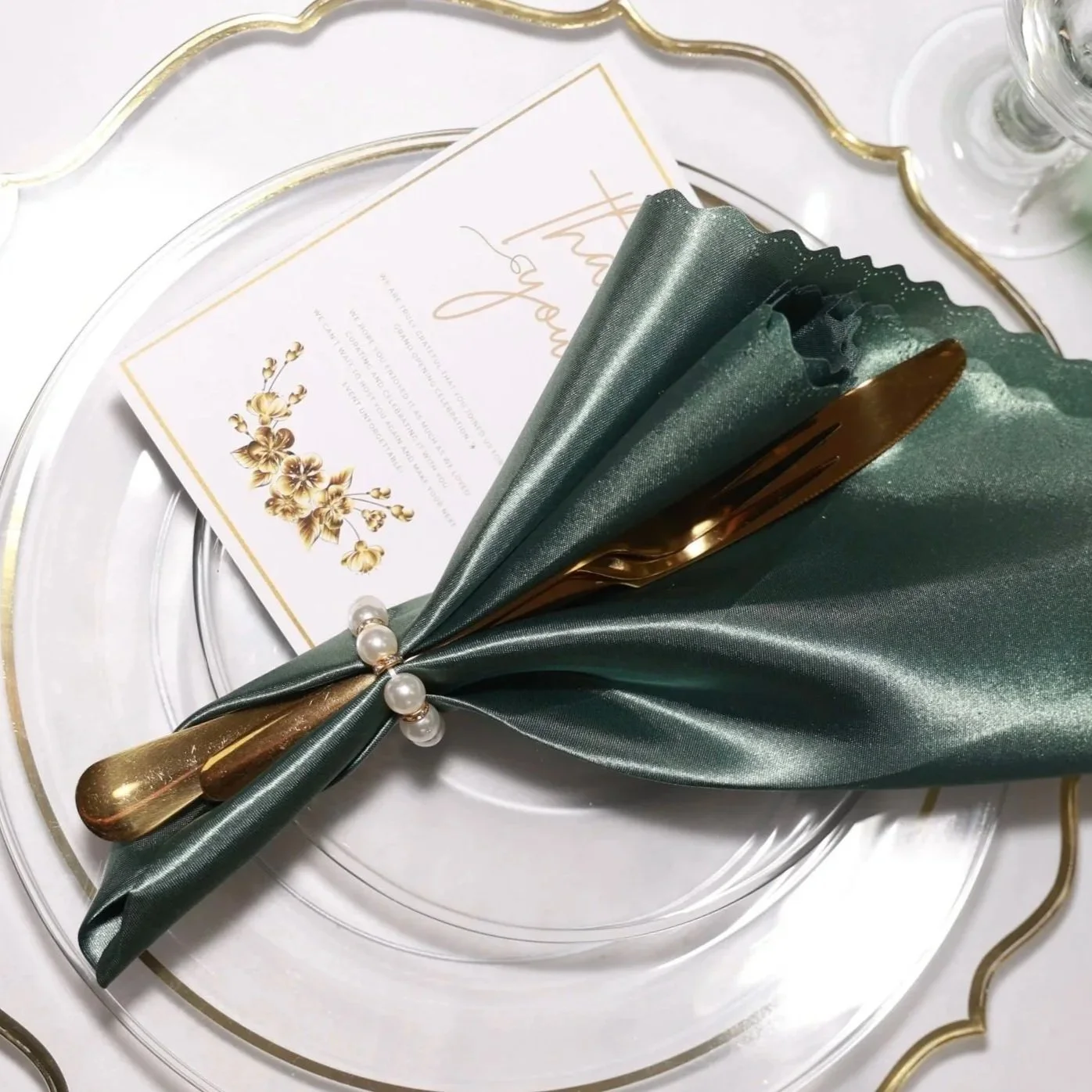 Elegant place setting with a gold-handled knife wrapped in green satin and lace, decorated with pearl accents, on a white plate with gold trim. A card with gold floral design is placed underneath.