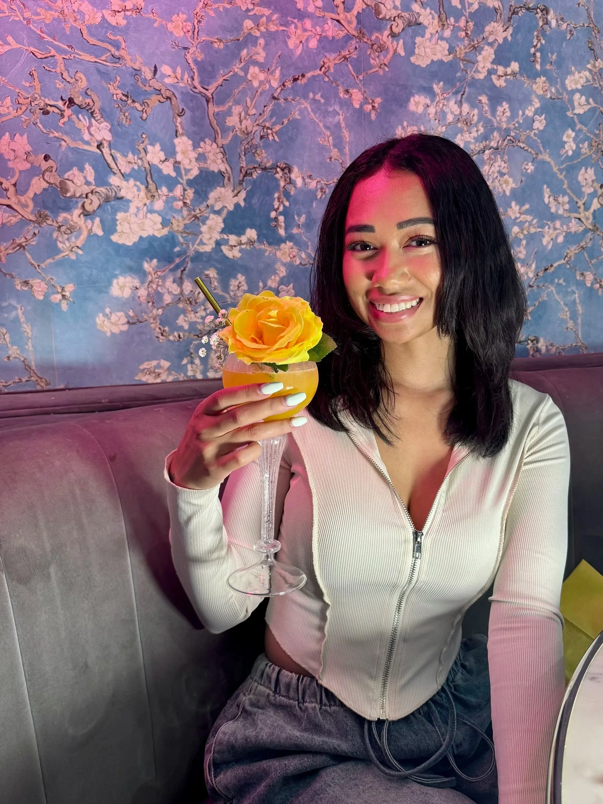 A young woman with shoulder-length black hair, smiling, holding a yellow cocktail garnished with a yellow flower and leaves, sitting on a gray upholstered bench against a blue floral wallpaper background.