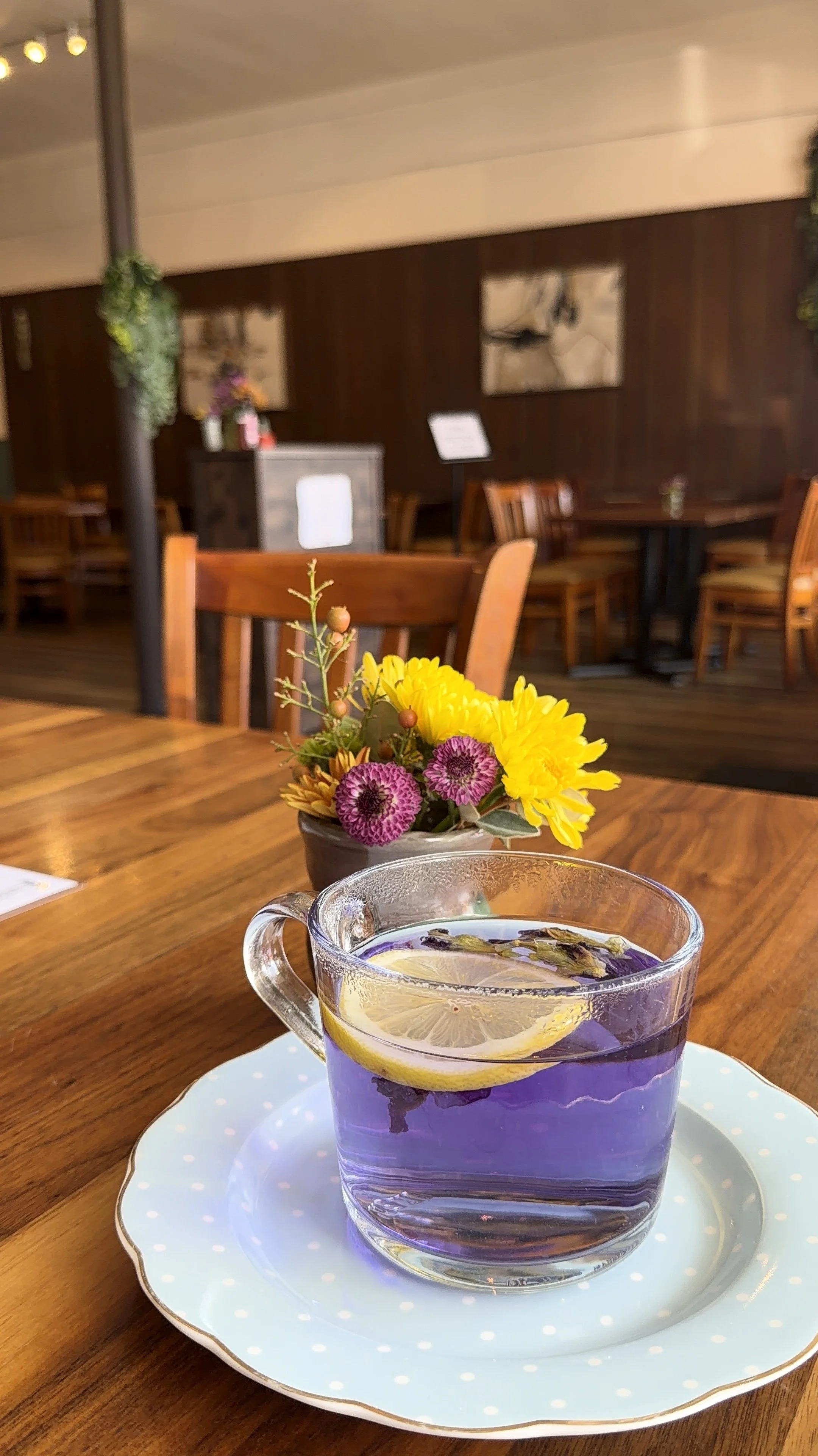 A glass cup of lavender tea with lemon slices, on a saucer, next to a small flower arrangement in a brown pot on a wooden table inside a cozy cafe or restaurant.