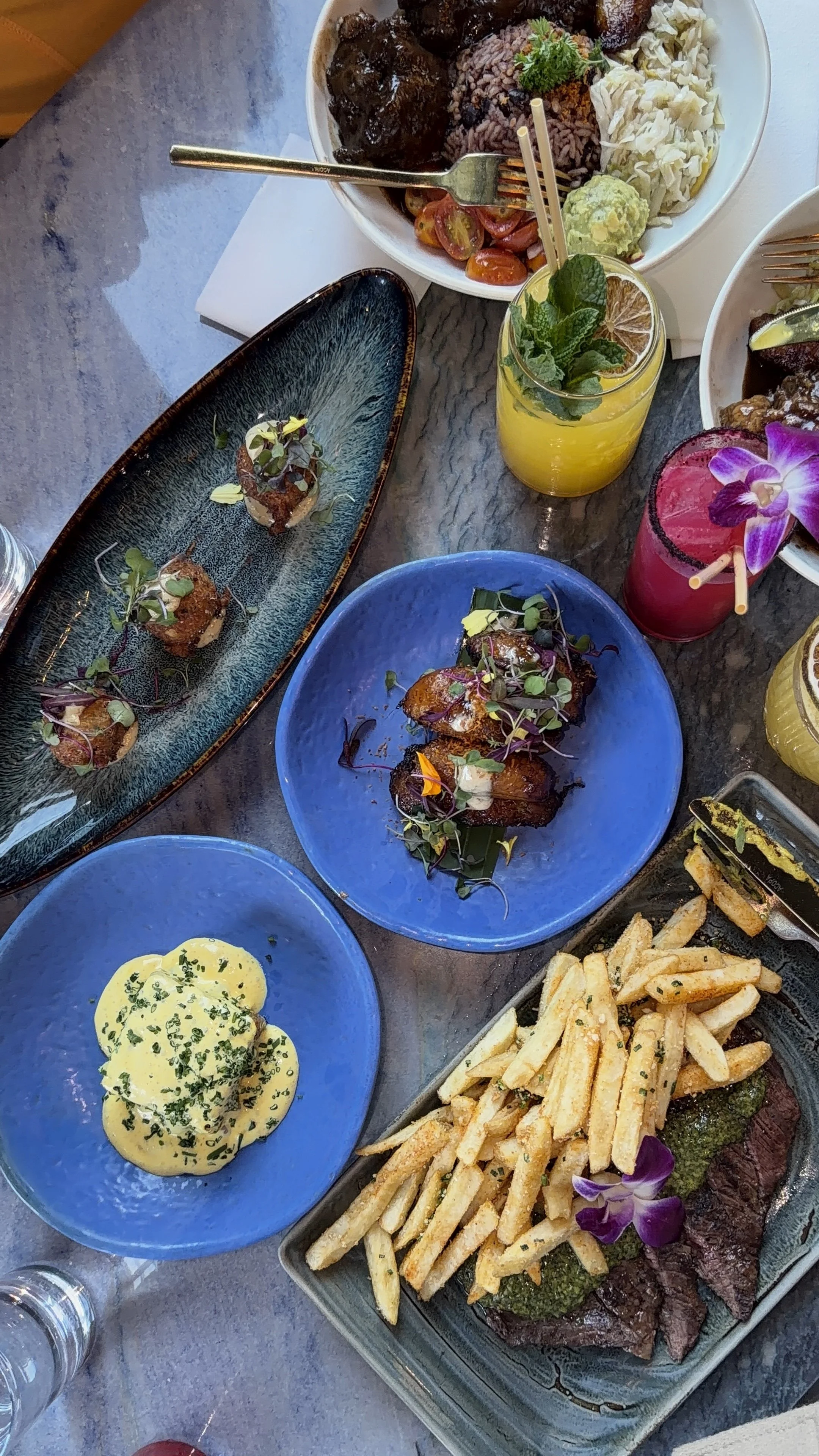 Various dishes including fries with steak, grilled meats with microgreens, rice, and vegetables, along with colorful beverages, on a table.
