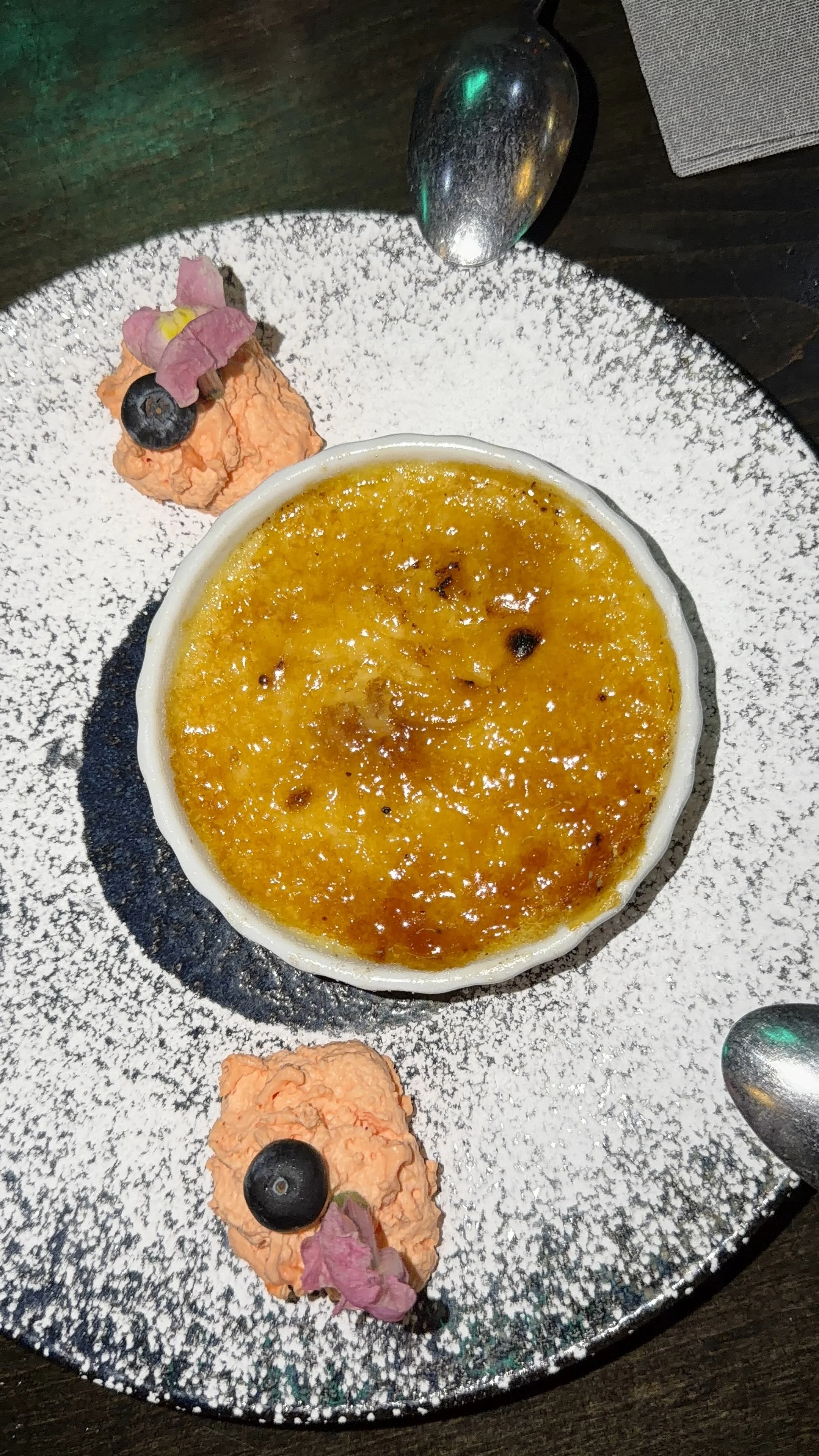 A dessert of crème brûlée with caramelized sugar top, garnished with blueberries, pink flower-shaped decorations, and a dollop of whipped cream with a blueberry on top, served on a plate with powdered sugar.